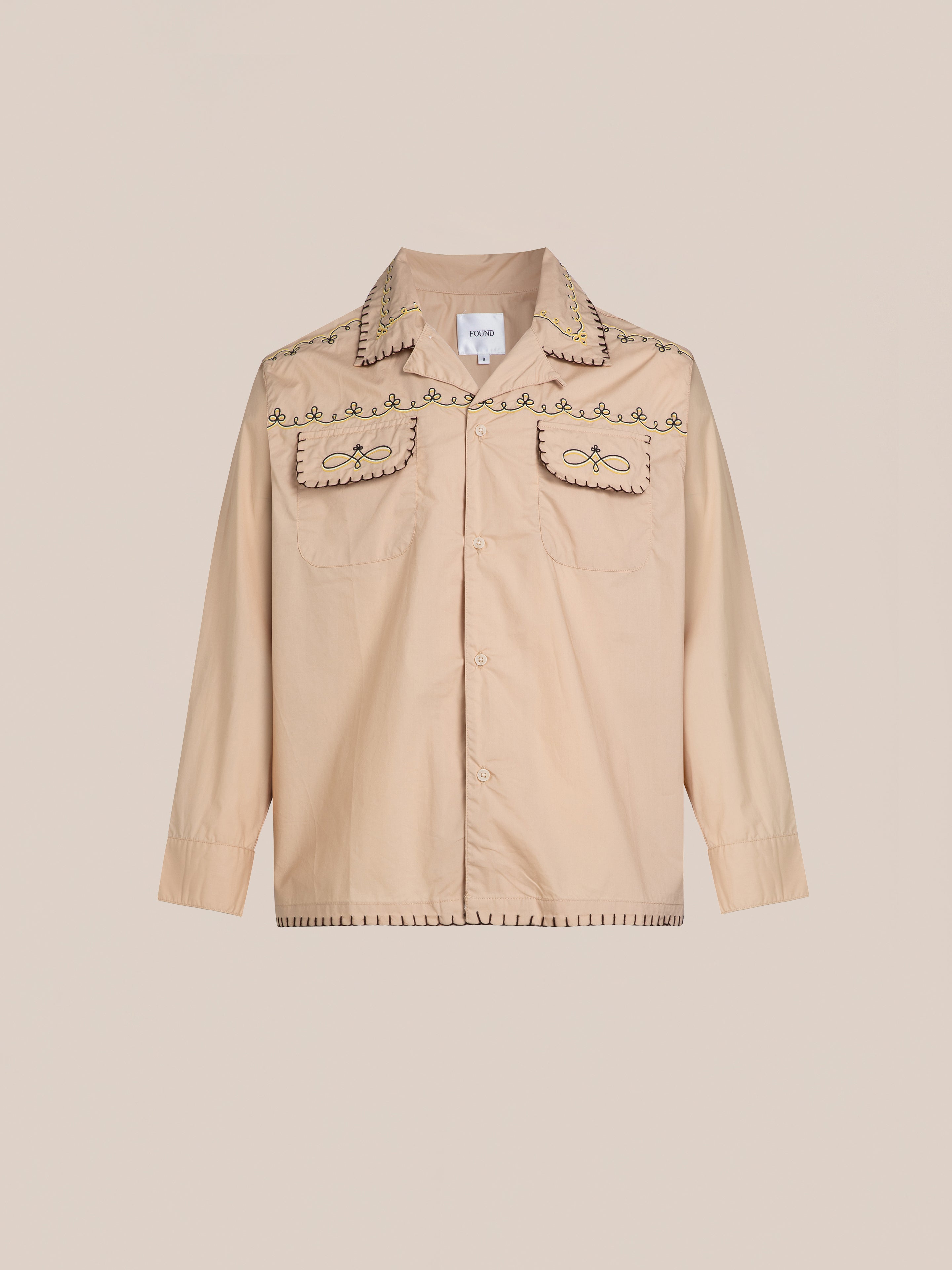 The Embroidered Western Button Up by FOUND is a beige shirt featuring intricate embroidered patterns on the collar, chest pockets, and hem.