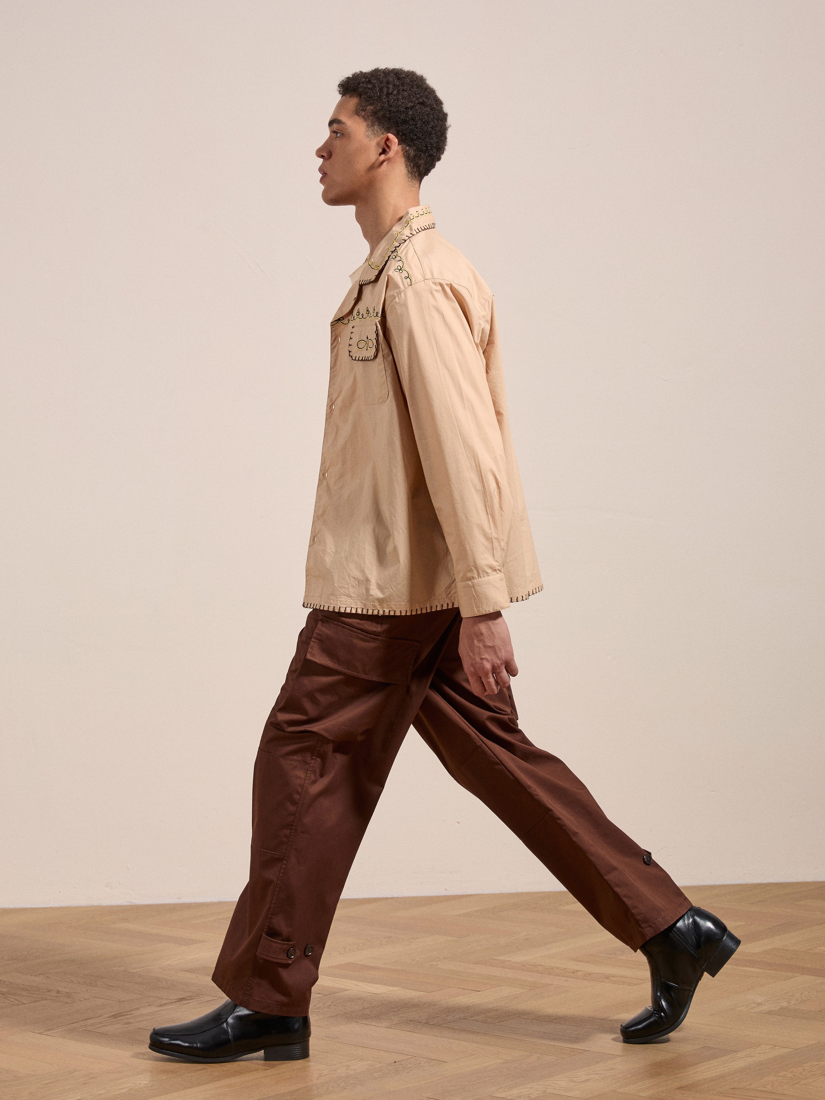 A person is walking sideways in an Embroidered Western Button Up by FOUND, brown pants, and black shoes on a wood floor against a plain background.