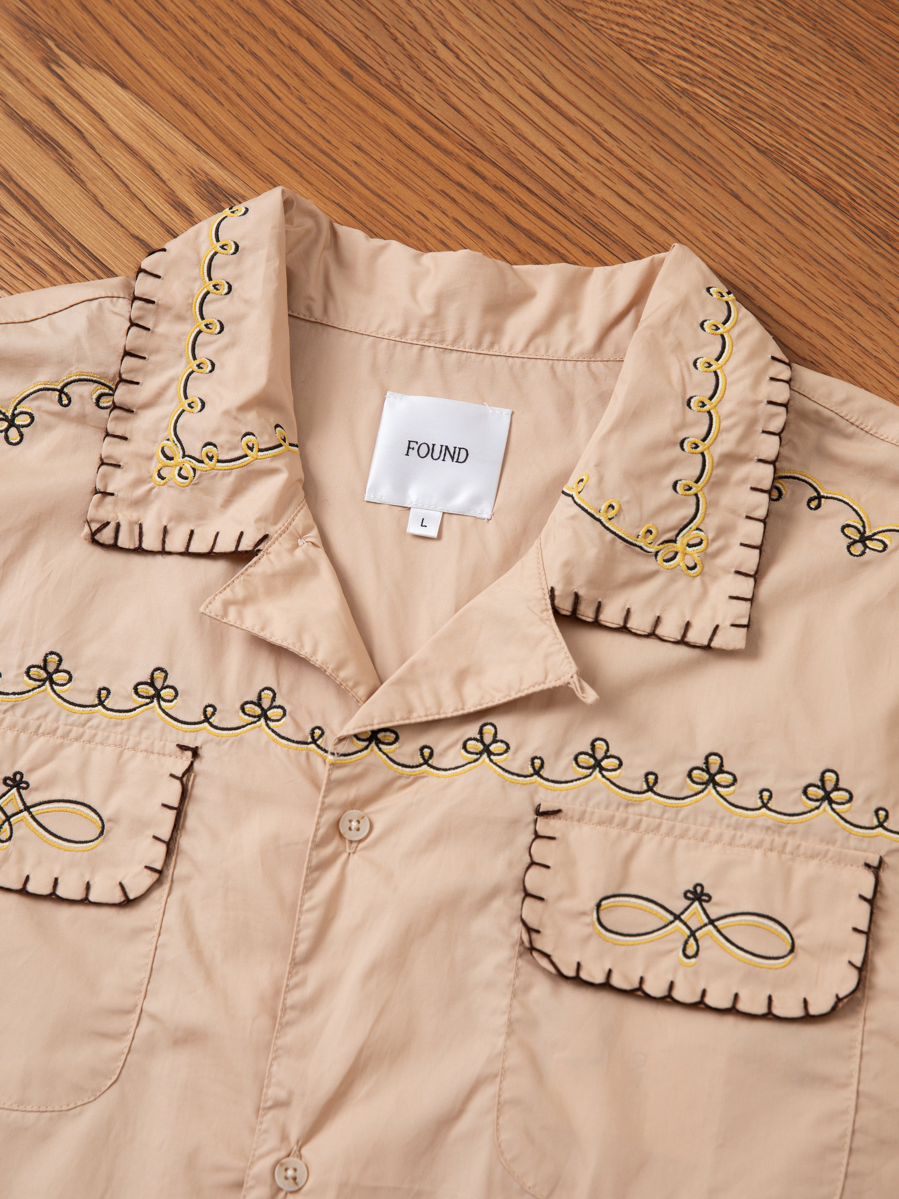 Displayed on a wooden surface is the Embroidered Western Button Up, a beige collared shirt adorned with decorative black and yellow embroidery on the collar, pockets, and front. Inside, a label reads "FOUND" and "L".