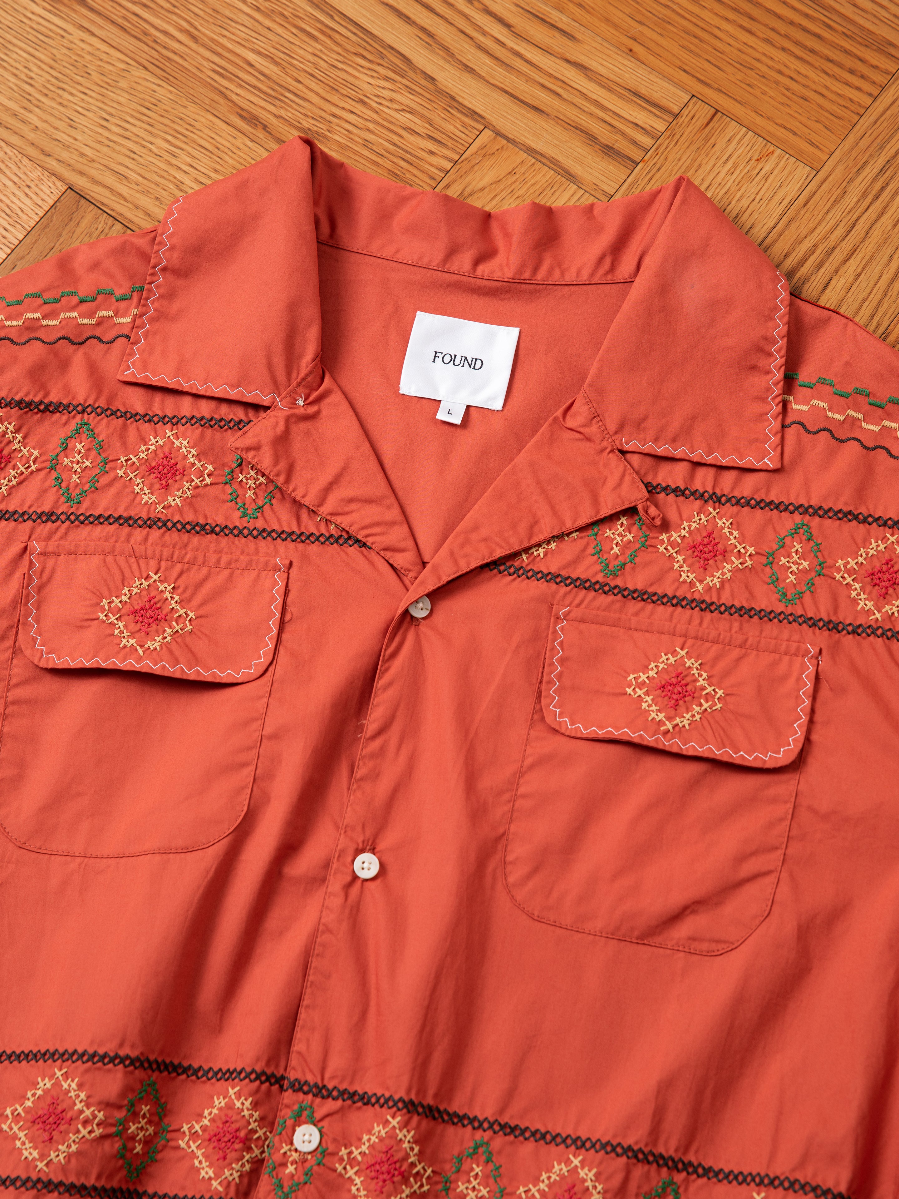 An Embroidered Western Button Up shirt, coral-colored with two front pockets and intricate embroidery patterns, is displayed on a wooden floor. A tag inside the collar reads "FOUND".