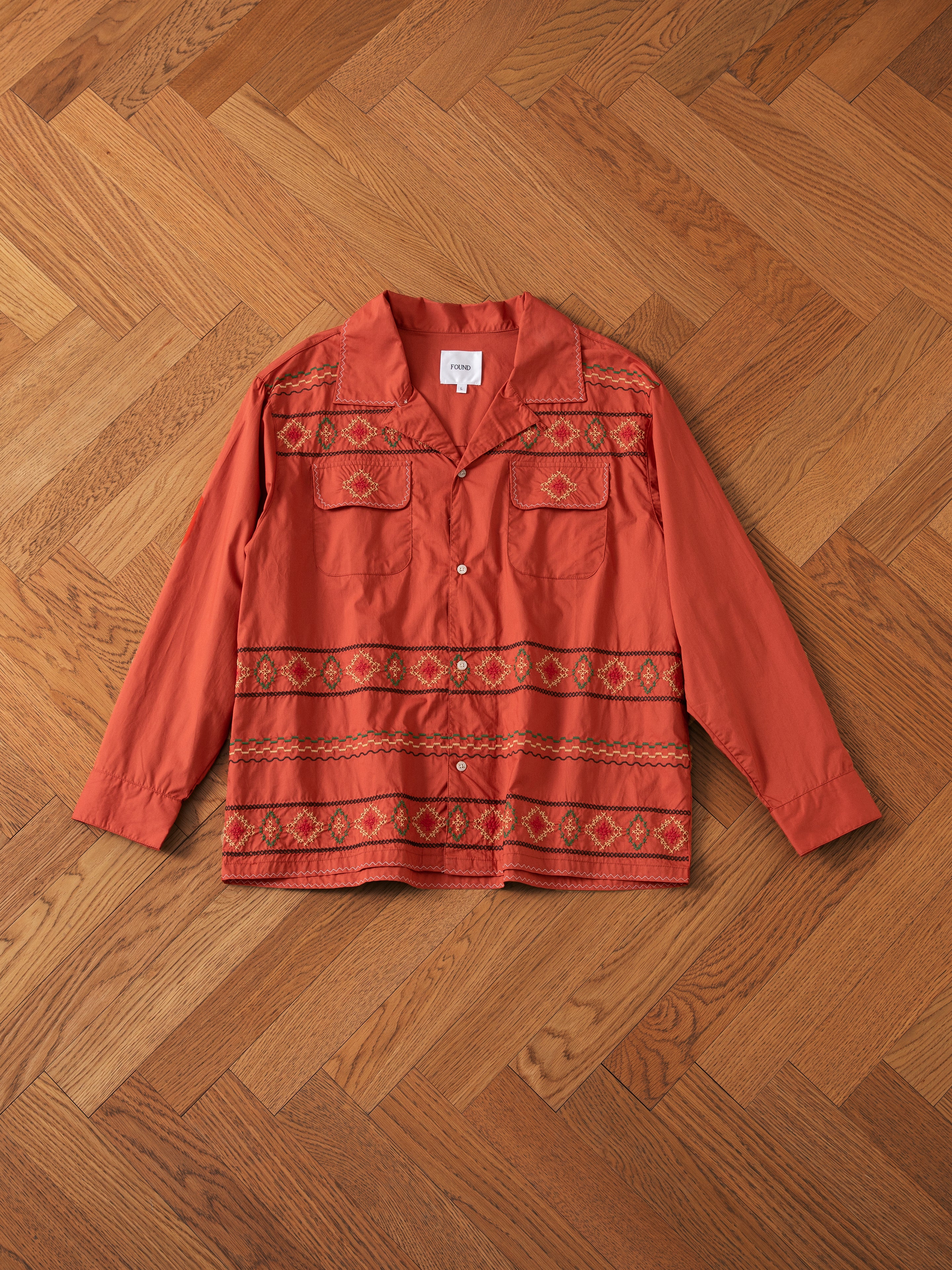 The FOUND Embroidered Western Button Up, a long-sleeved shirt in burnt orange with intricate red and brown embroidered patterns, is laid flat on a wooden herringbone floor.