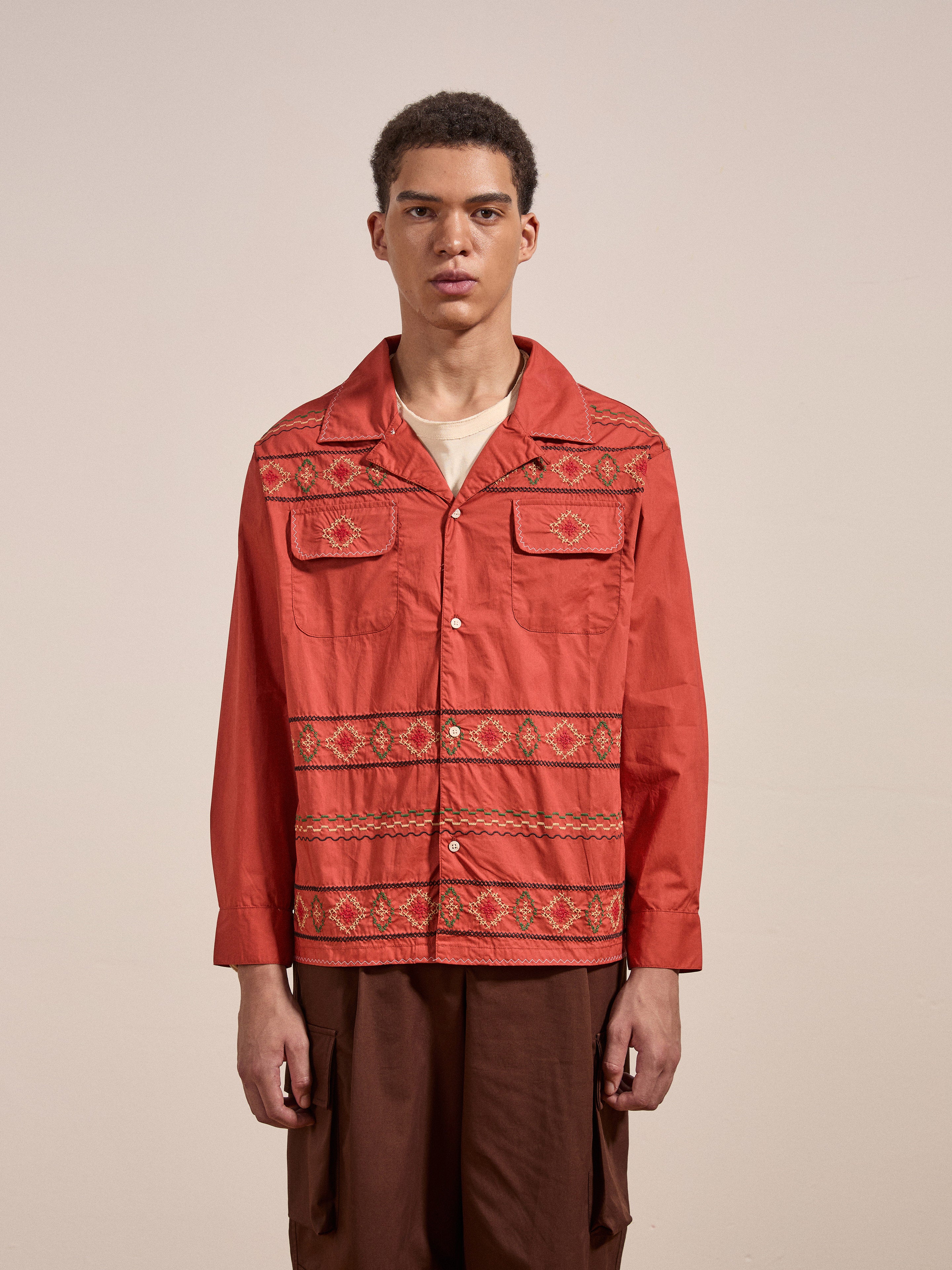 A person stands facing forward wearing FOUND's Embroidered Western Button Up, a red shirt with two chest pockets and intricate embroidered patterns, paired with brown trousers. The background is plain off-white.