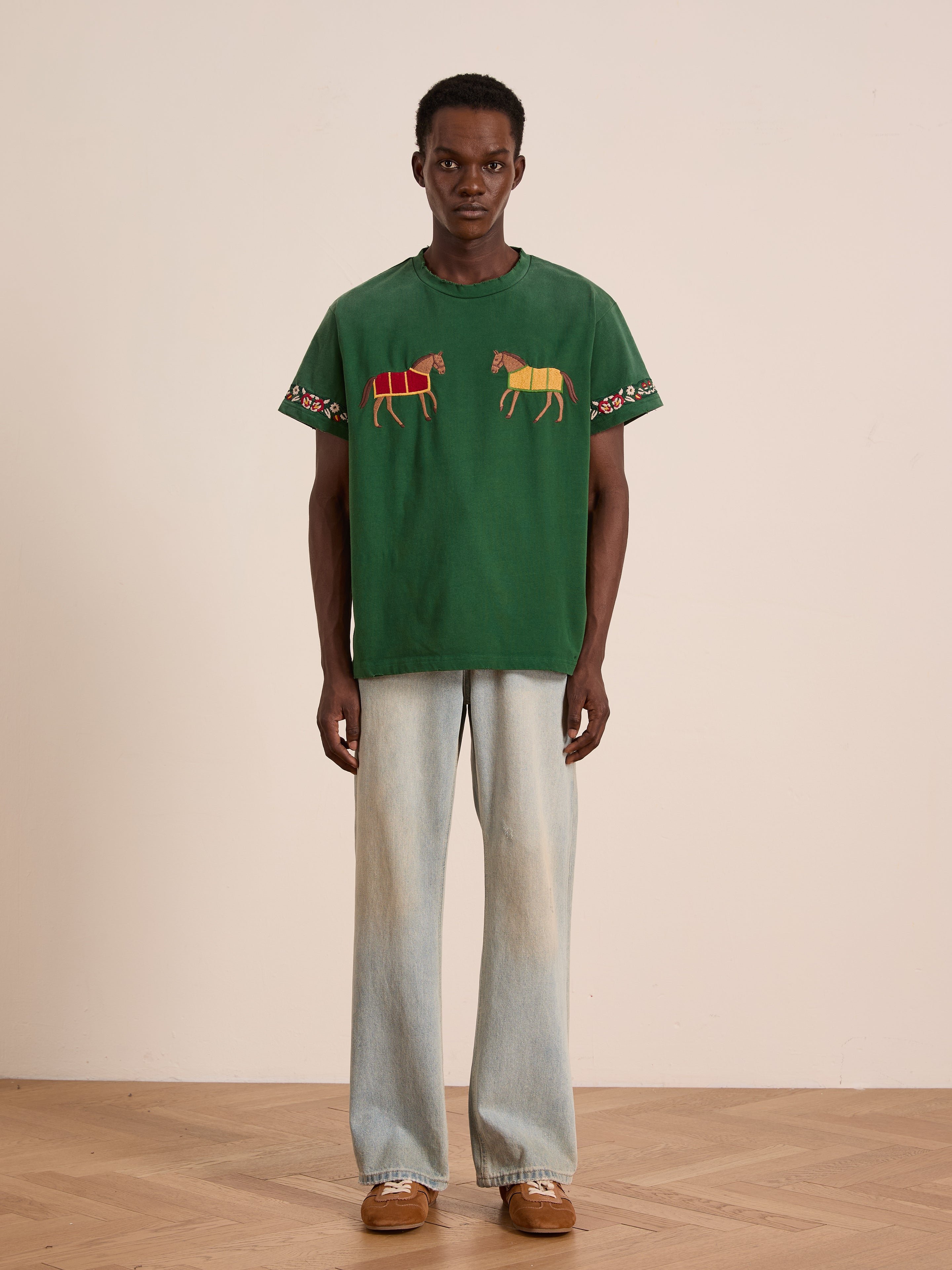 A person stands facing forward against a plain background, wearing the FOUND Horse Equine Tee with embroidered horses on the sleeves, paired with light blue jeans and brown shoes.