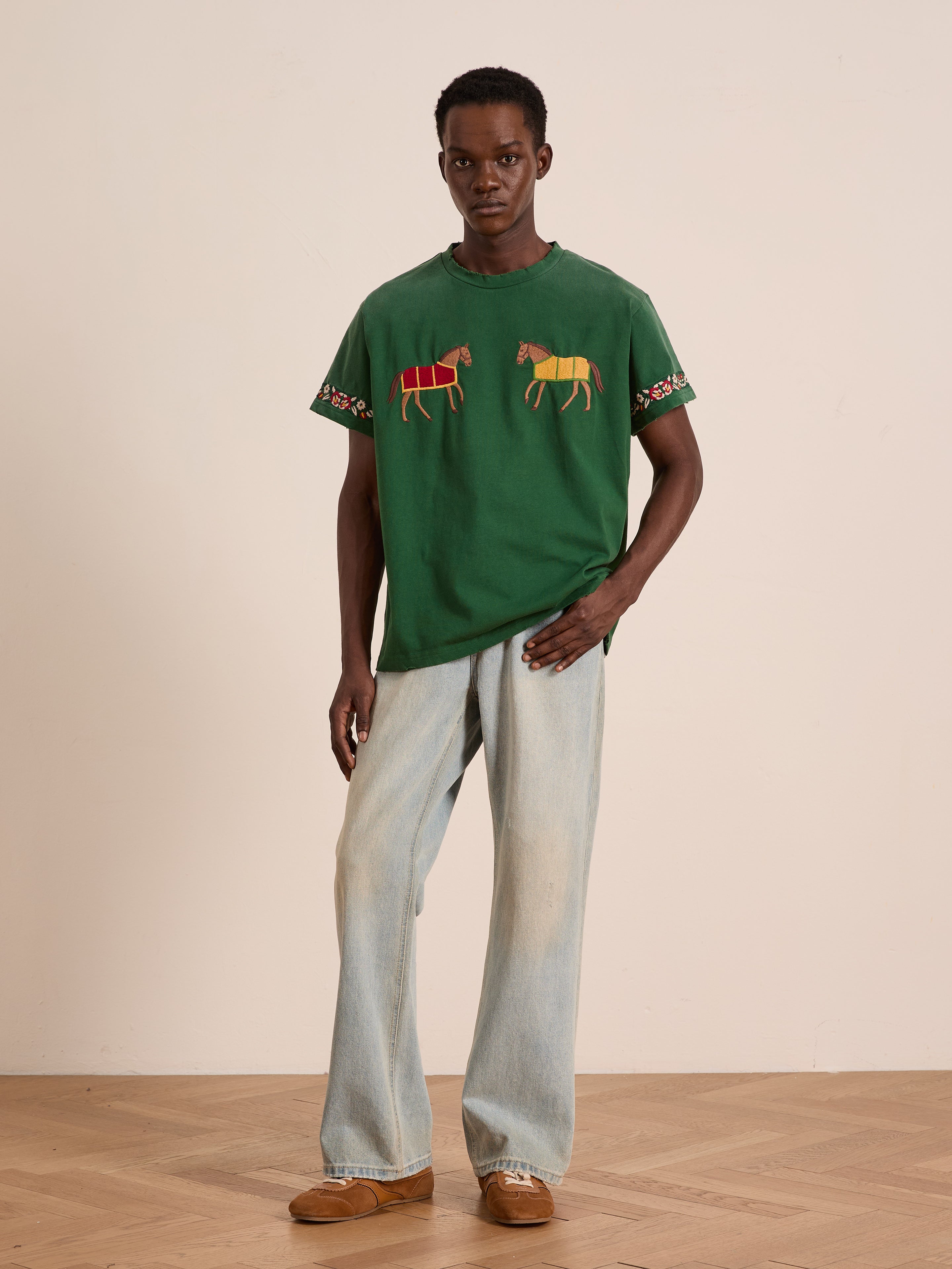 A person stands indoors wearing the FOUND Horse Equine Tee, featuring embroidered horses, styled with light-wash wide-leg jeans and brown shoes.