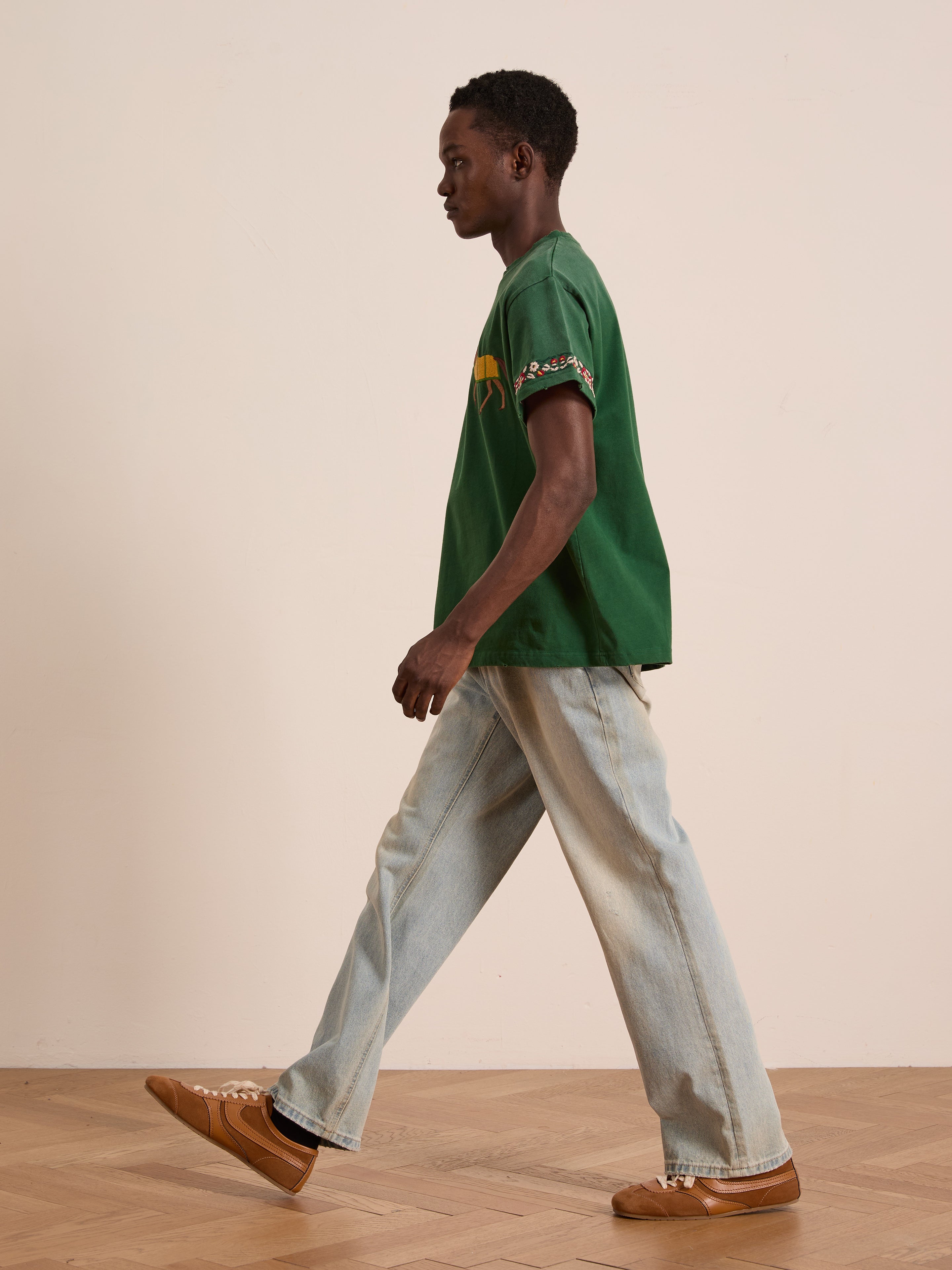 A man wears the FOUND Horse Equine Tee, paired with light blue jeans and brown sneakers, as he walks indoors on a wooden floor beside a plain light-colored wall.