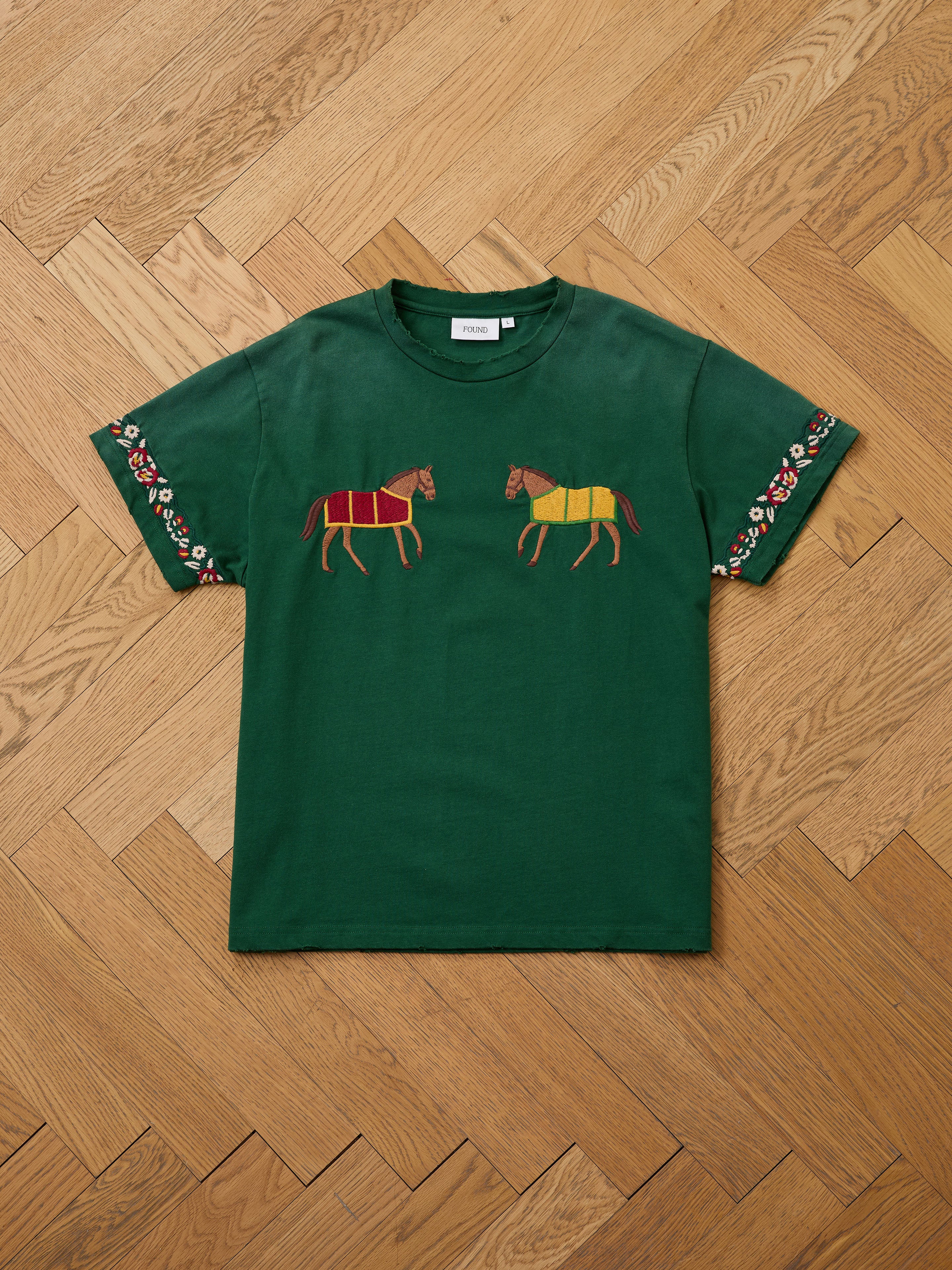 FOUND Horse Equine Tee: green short-sleeve shirt with embroidered horses on the chest and floral sleeve accents, shown laid flat on a herringbone wooden floor.