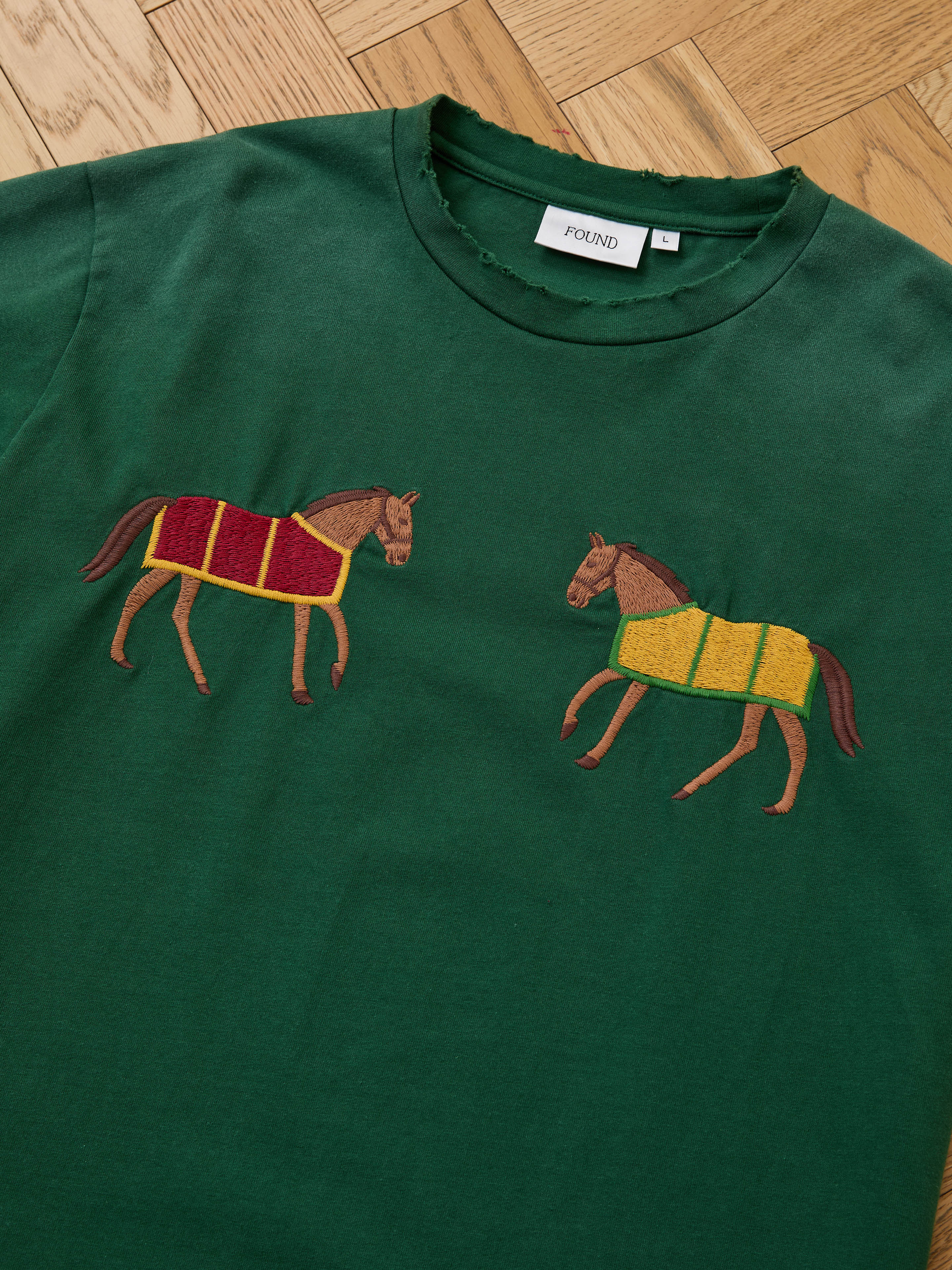 The FOUND Horse Equine Tee, a green T-shirt featuring embroidered horses with jockeys in red/yellow and yellow/green uniforms, is displayed flat on a wooden floor.