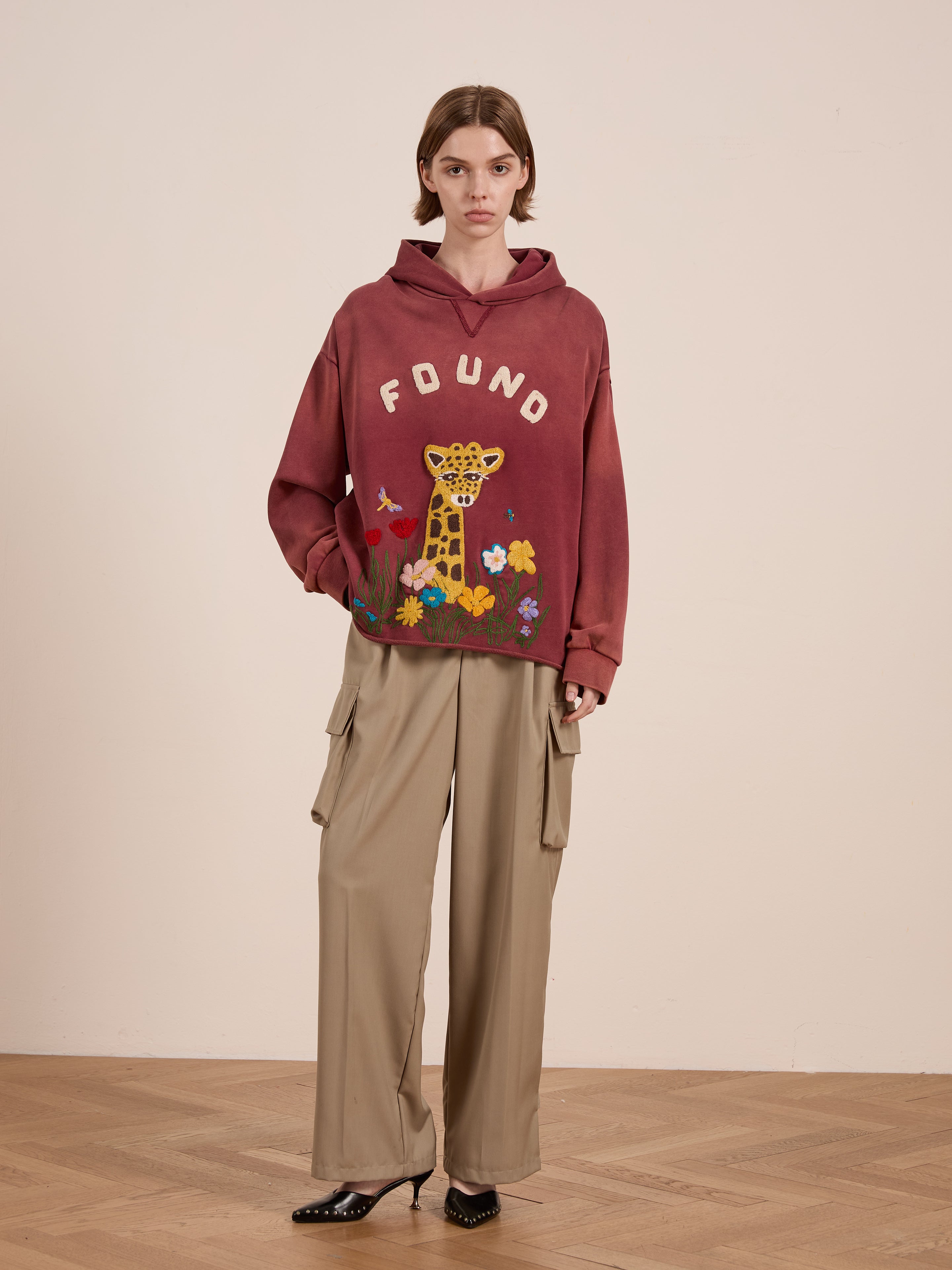 A person in a FOUND Embroidered Wildlife Hoodie, with giraffe and flower detailing, paired with beige cargo pants stands on a wooden floor. The hoodie’s unisex relaxed fit enhances style, comfort, and versatility.