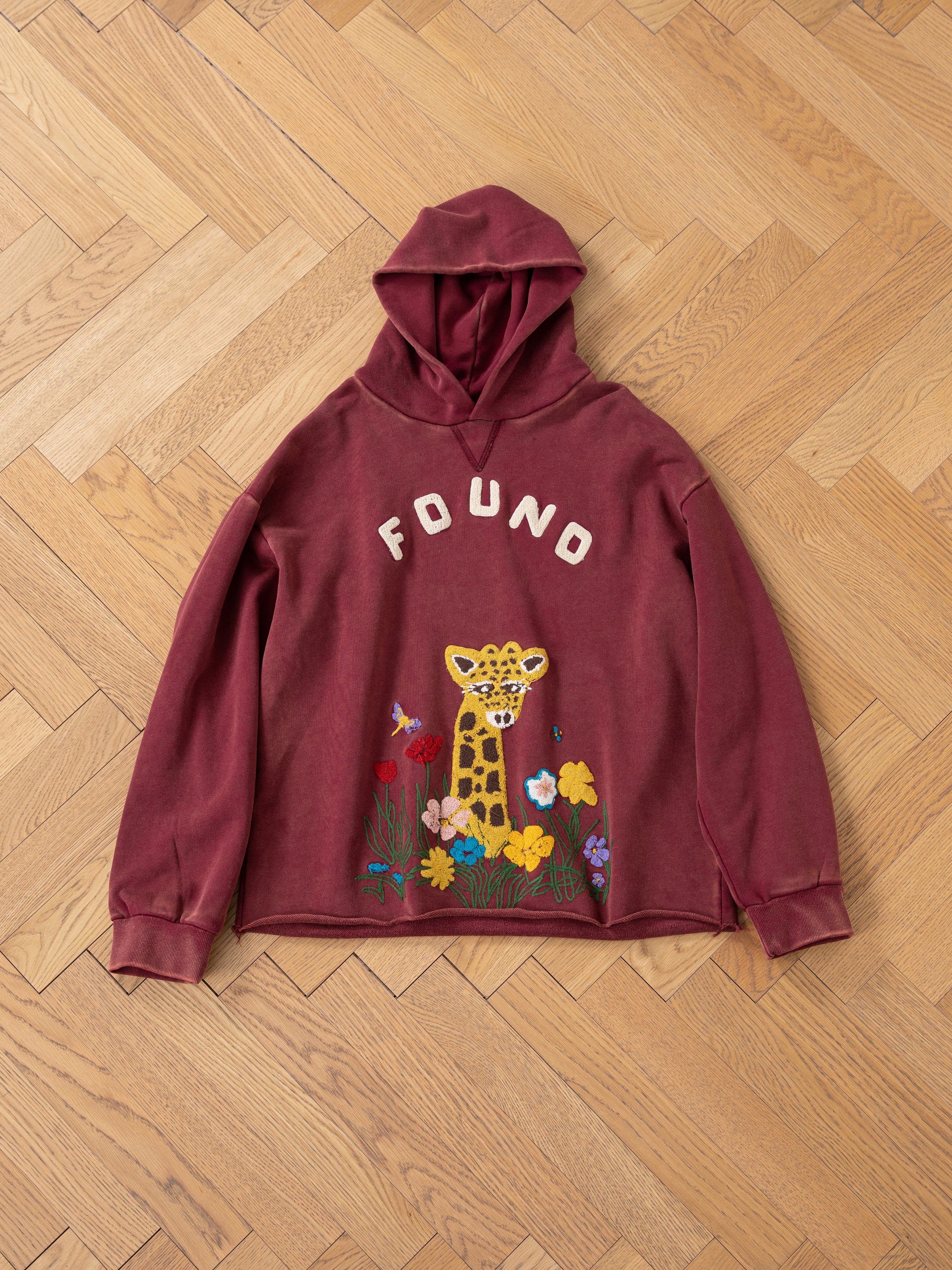 The FOUND Embroidered Wildlife Hoodie is a maroon, unisex relaxed fit featuring "FOUND" text and a giraffe amid colorful flowers with intricate wildlife embroidery on the front, artfully displayed on a wooden herringbone floor.