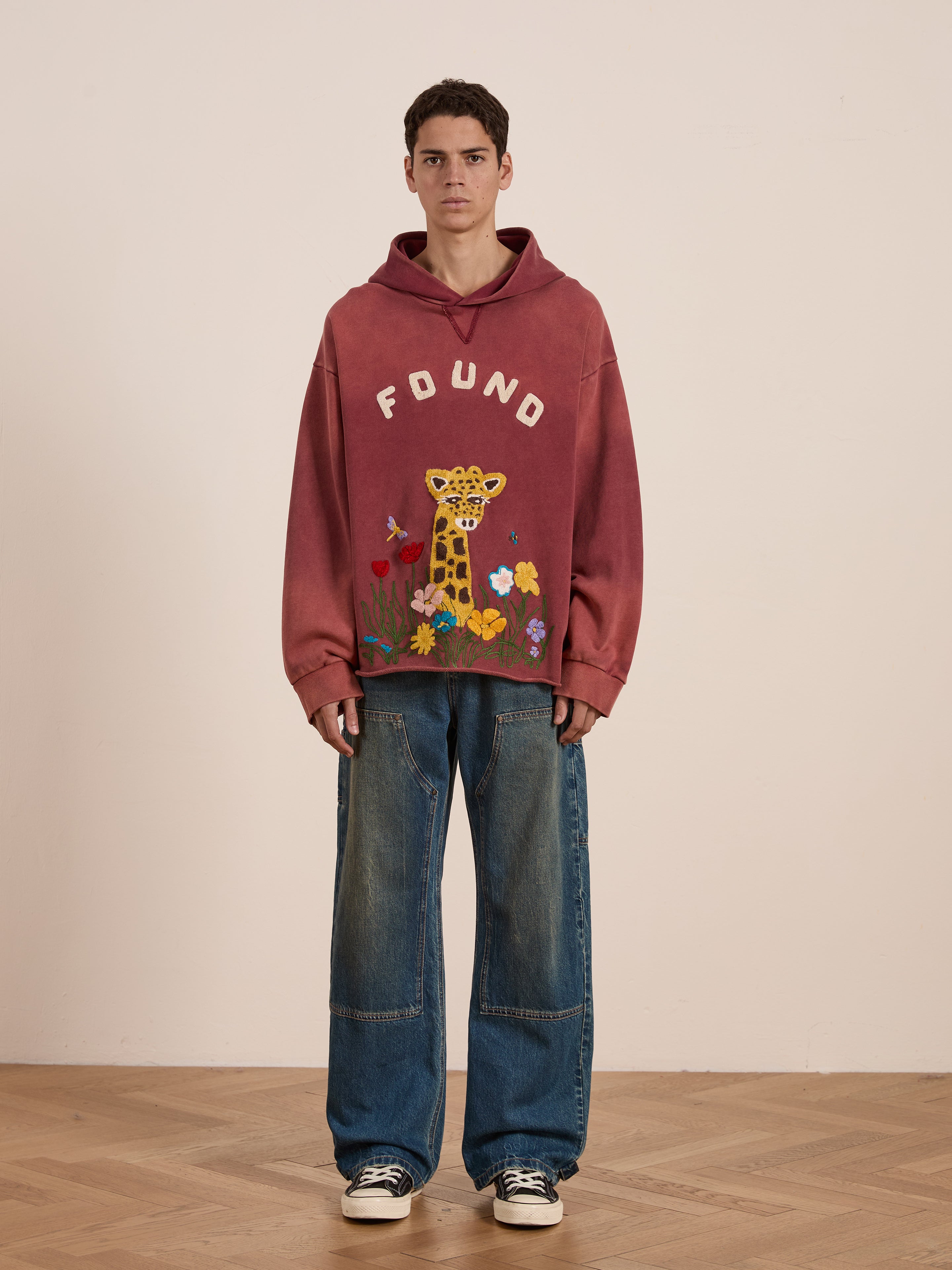 A person stands on a wooden floor wearing the Embroidered Wildlife Hoodie by FOUND, featuring pink fabric with a giraffe design. Paired with wide-legged jeans and sneakers, the unisex relaxed fit hoodie adds unique flair to the casual look.