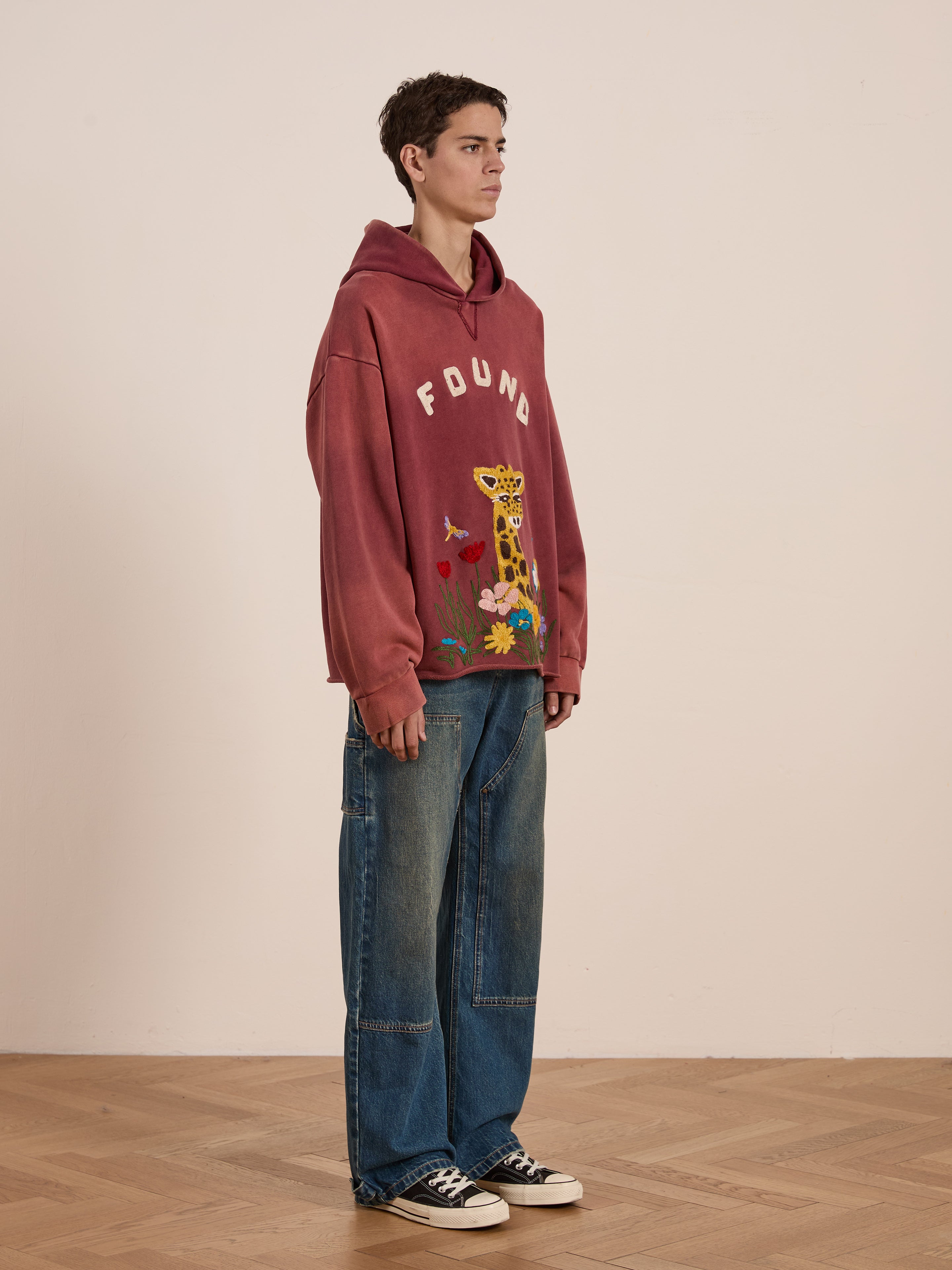 A person stands sideways in a room, sporting the Embroidered Wildlife Hoodie by FOUND featuring animal print and "FOUND" embroidery, paired with baggy jeans and black sneakers. The unisex relaxed fit offers a casual yet stylish vibe.