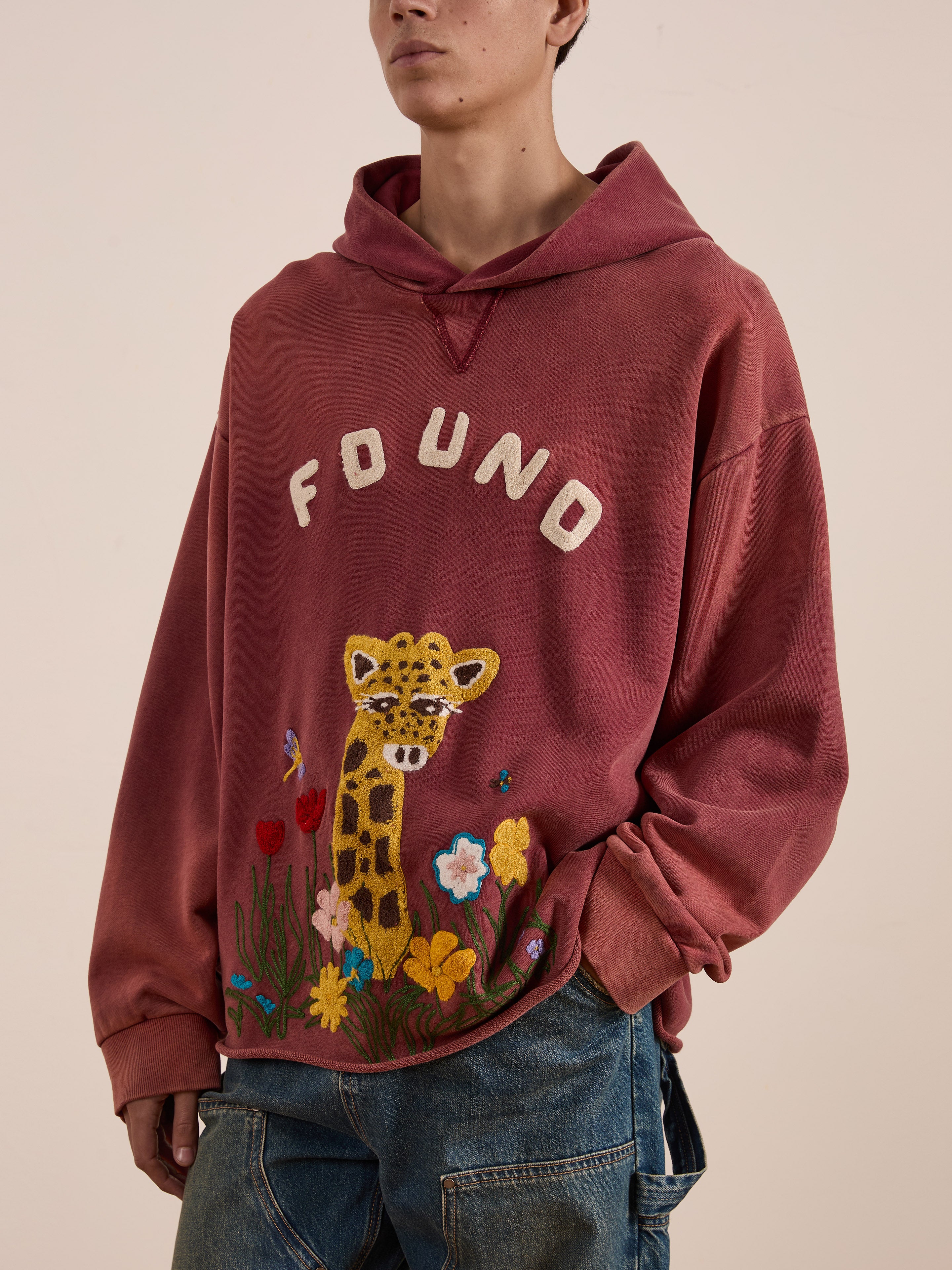 Someone is wearing the unisex relaxed fit Embroidered Wildlife Hoodie by FOUND, featuring "FOUND" text and a giraffe with colorful flowers in maroon. They’ve effortlessly paired it with blue jeans.