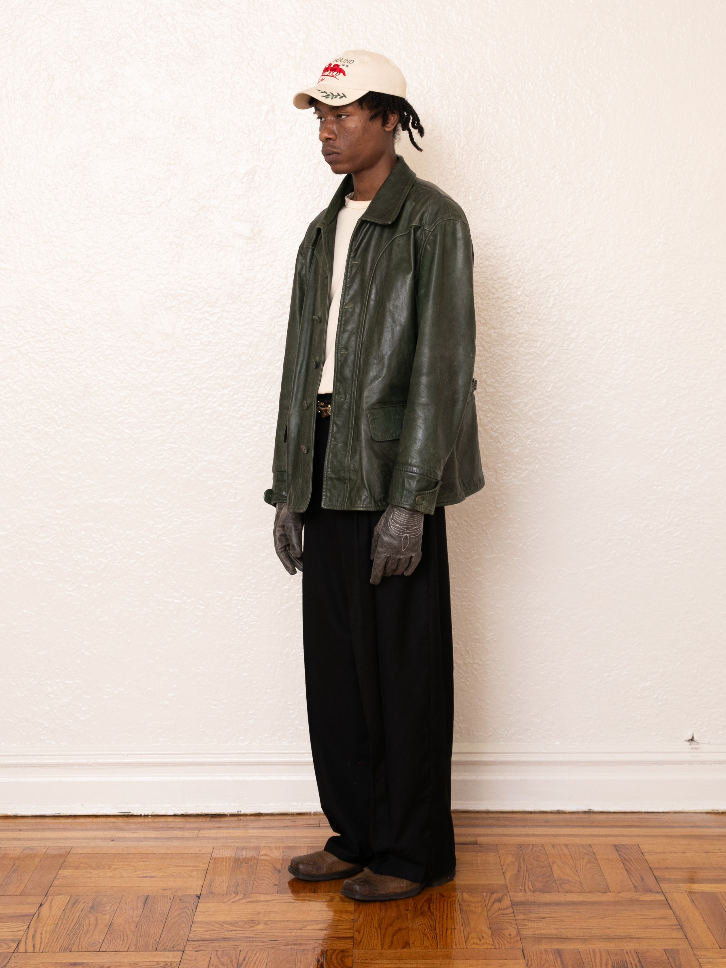 A person stands against a white wall wearing a FOUND Emerald Leather Jacket made from ethical materials, dark pants, gloves, and a white cap with a red logo.