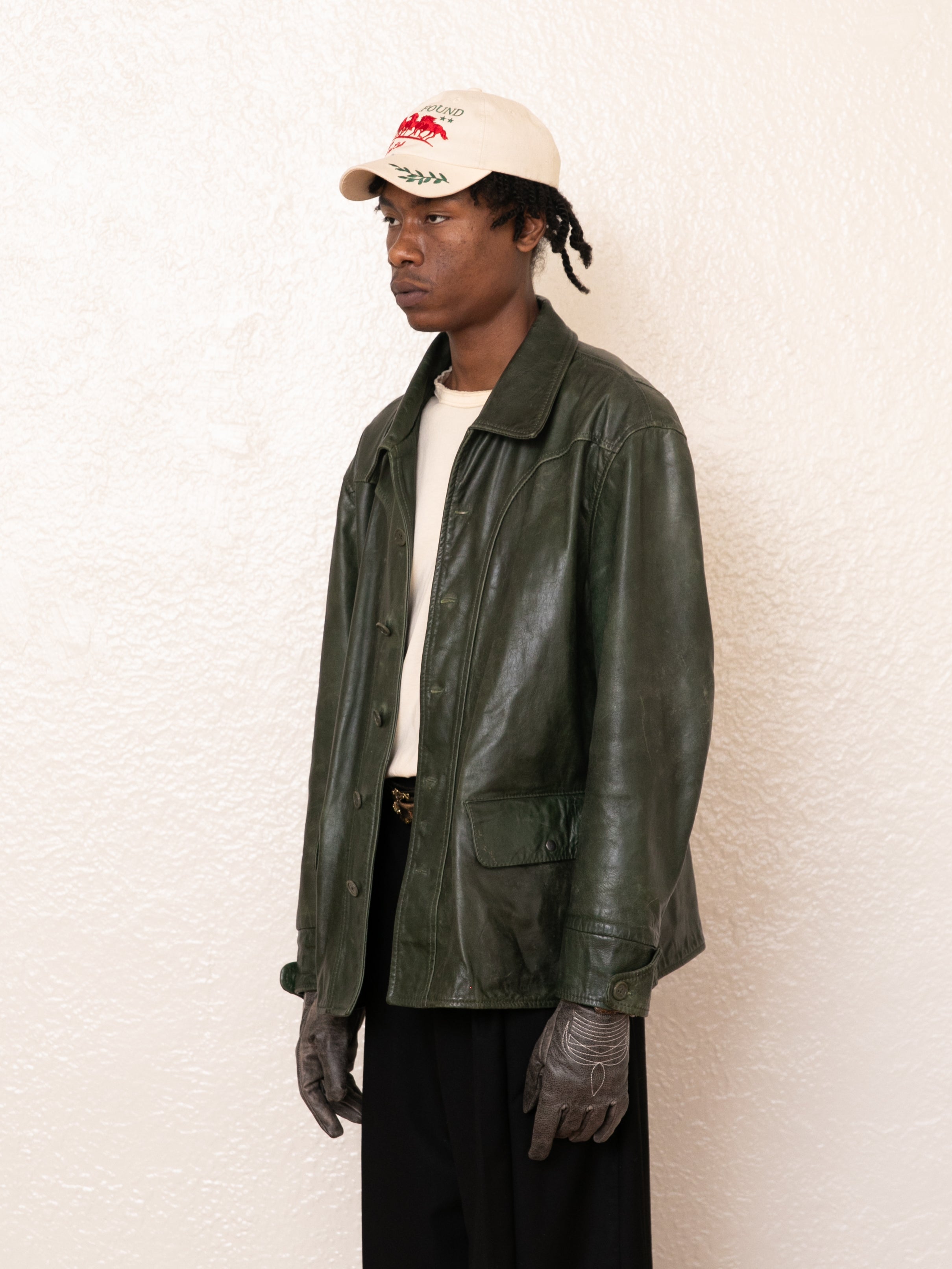 A person stands against a white textured wall, wearing a FOUND Emerald Leather Jacket made from ethically sourced materials, along with a beige cap and distressed gray gloves.