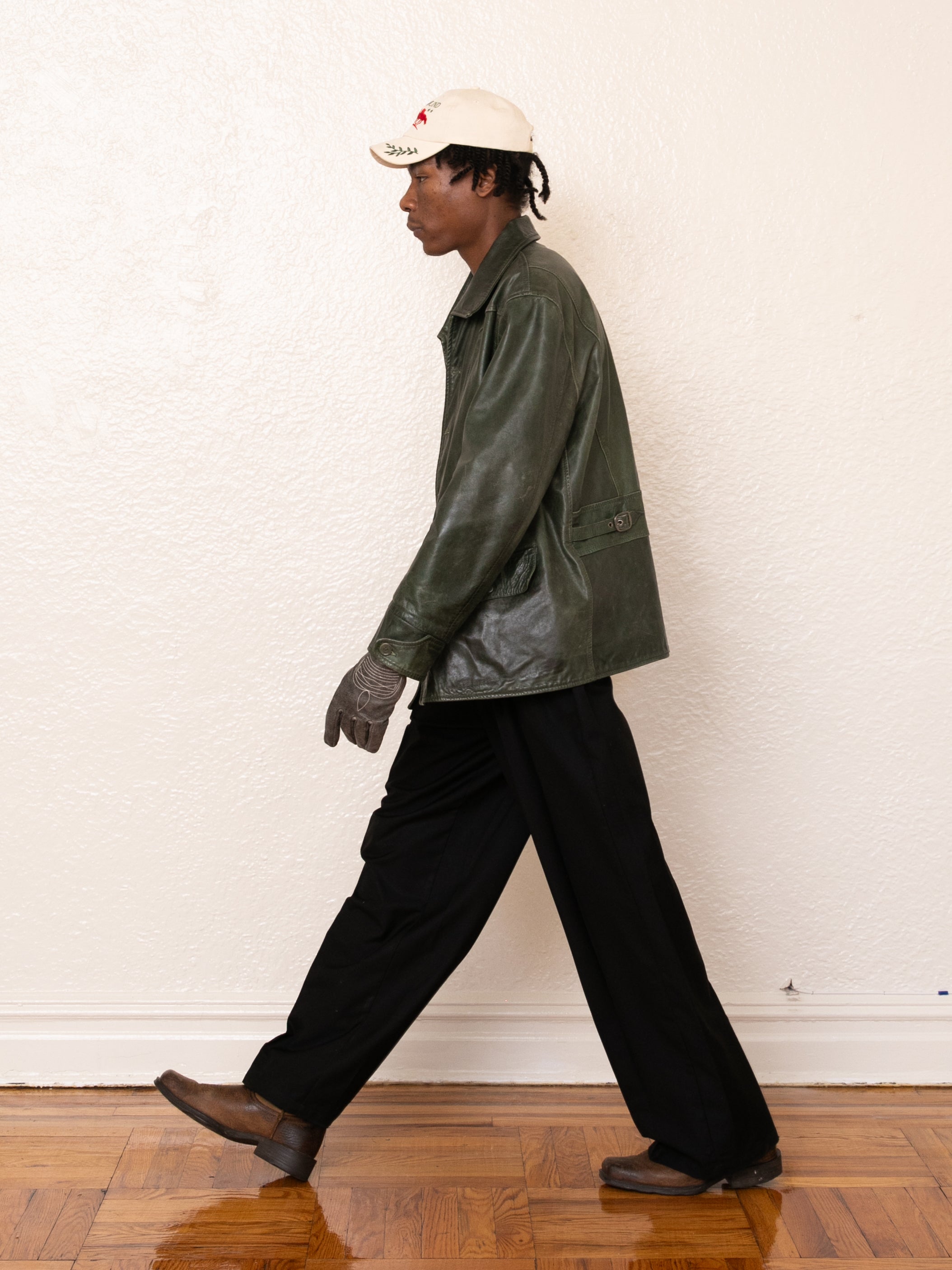 Dressed in a FOUND Emerald Leather Jacket made from ethically sourced materials, a person pairs it with black pants, brown shoes, gray gloves, and a white cap while walking against a plain white wall.