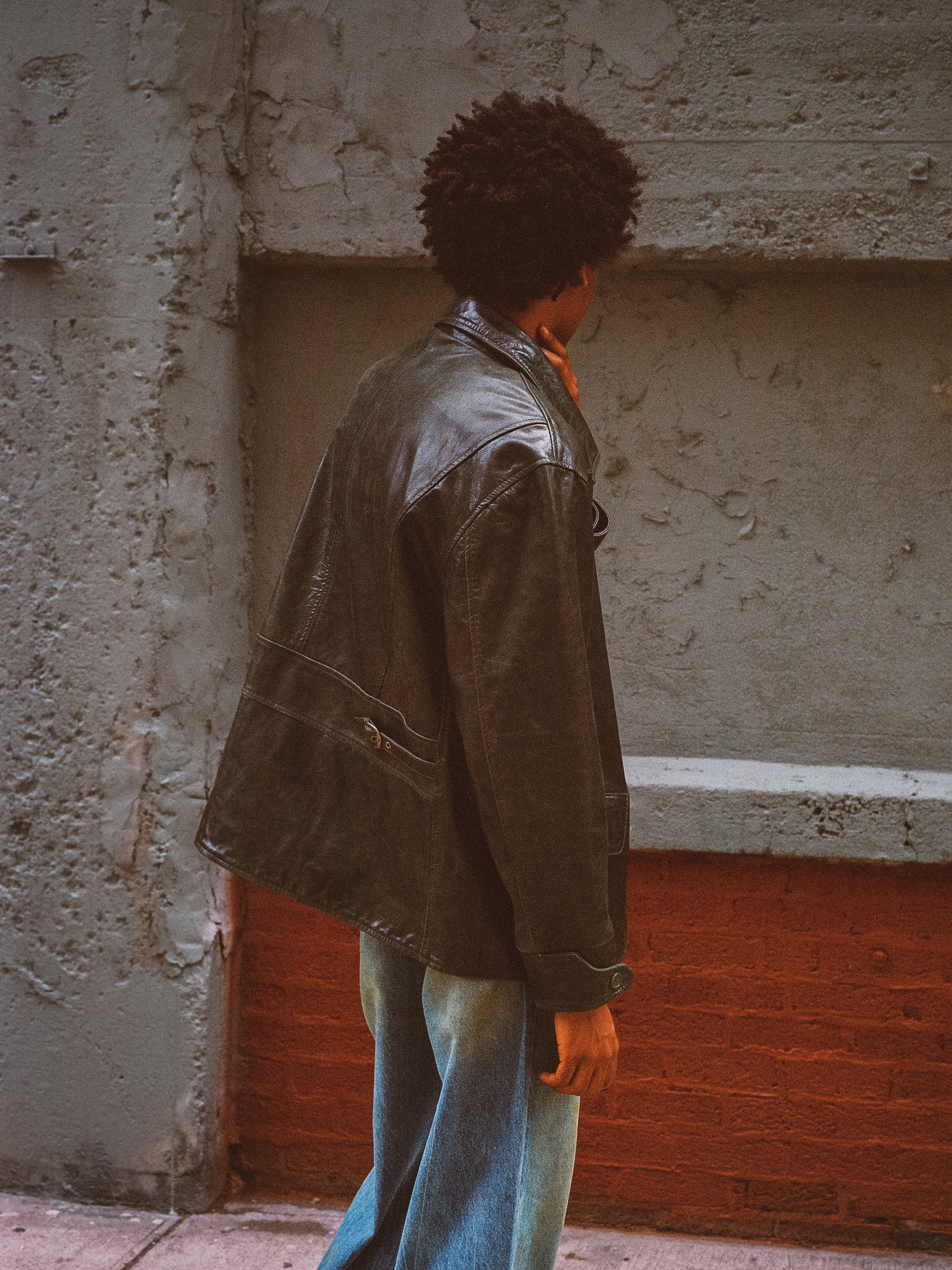 A person wearing an Emerald Leather Jacket by FOUND, crafted from ethically sourced materials, and jeans stands against a textured wall, looking away.