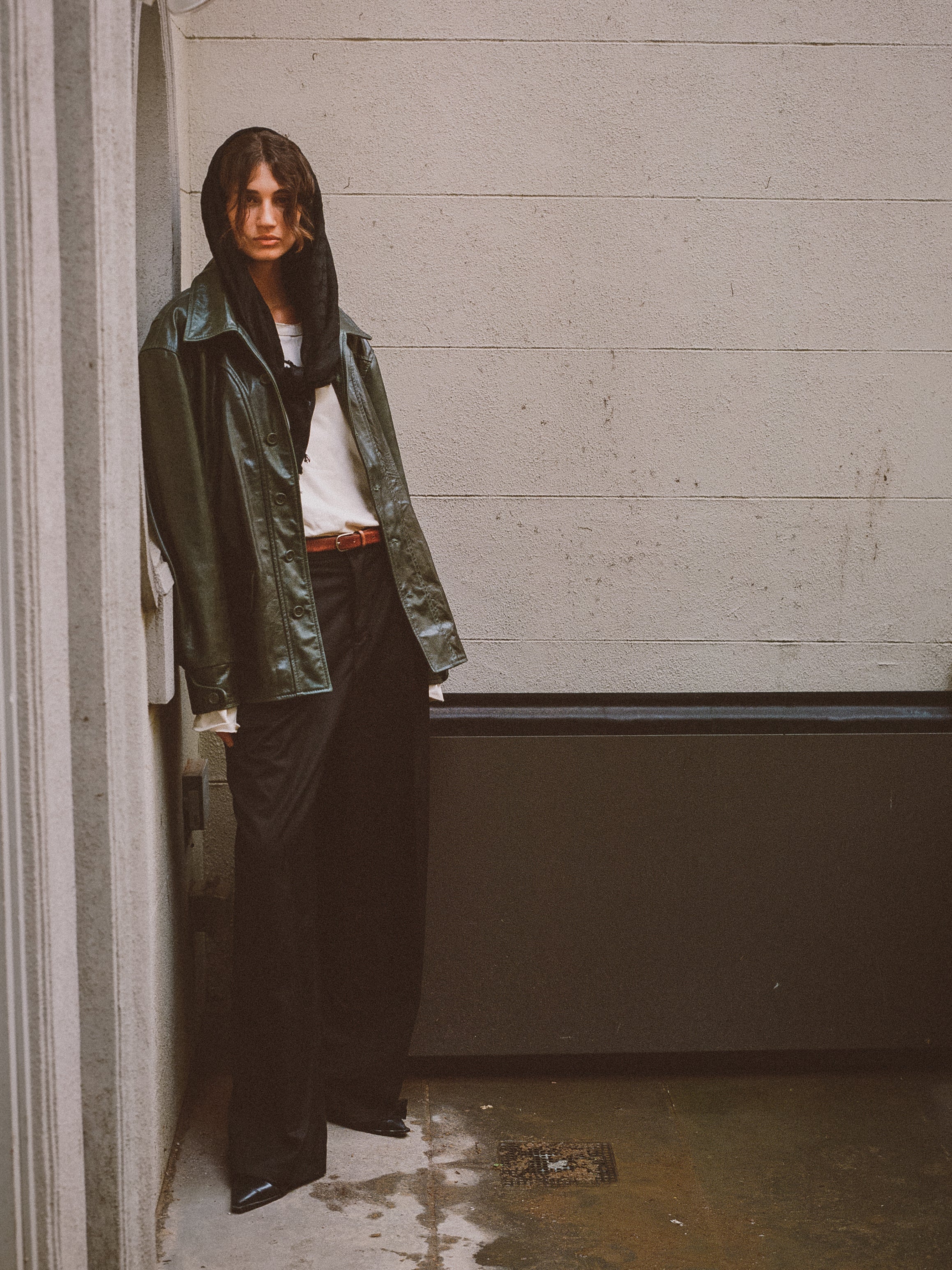 A person in a FOUND brand Emerald Leather Jacket, a white shirt, and black pants stands against a concrete wall in an alleyway.