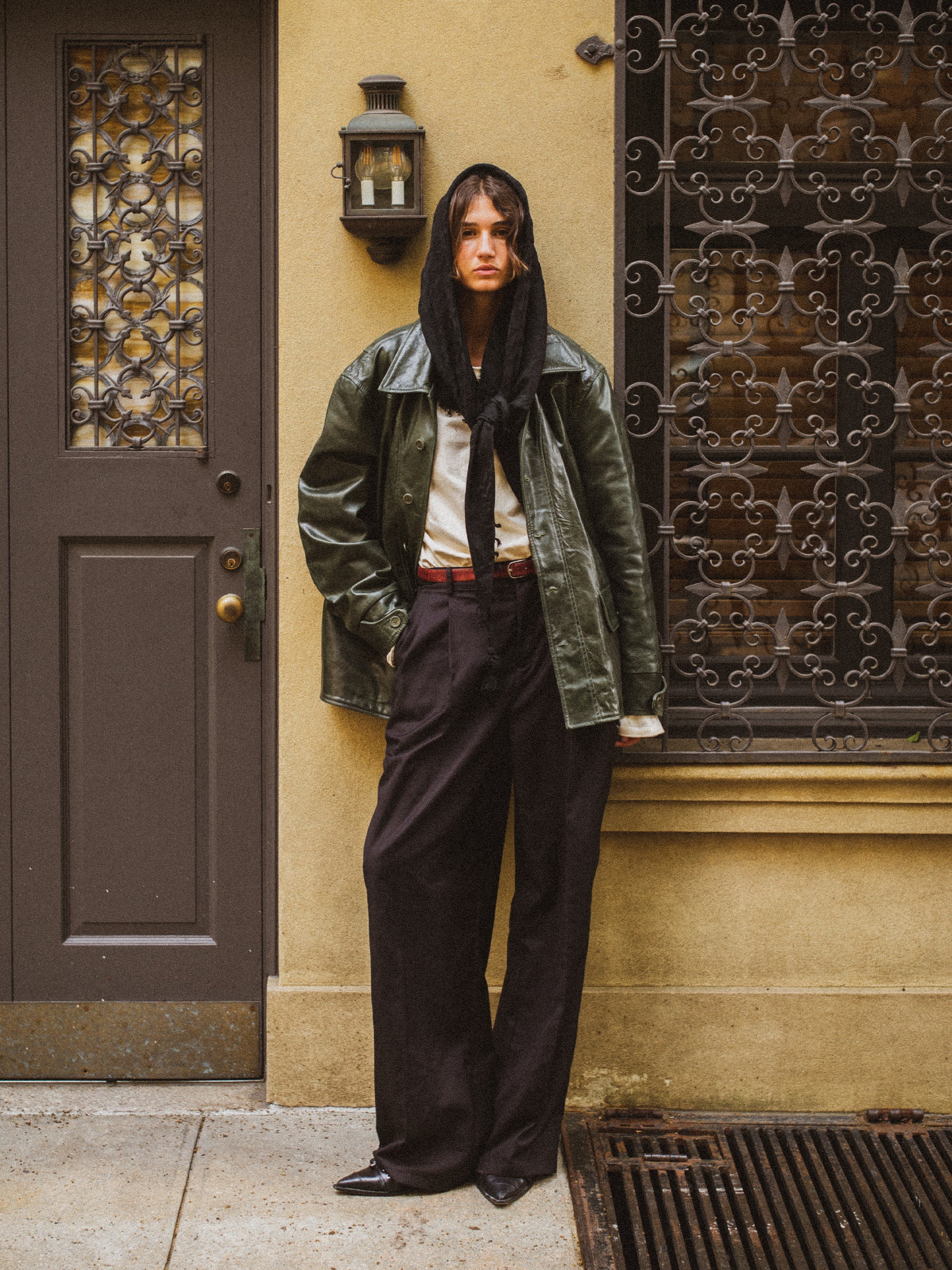 Wearing a FOUND Emerald Leather Jacket with a black scarf and pants, a person leans against a beige building with decorative metalwork and door, showcasing an eco-friendly blend of fashion and sustainability.