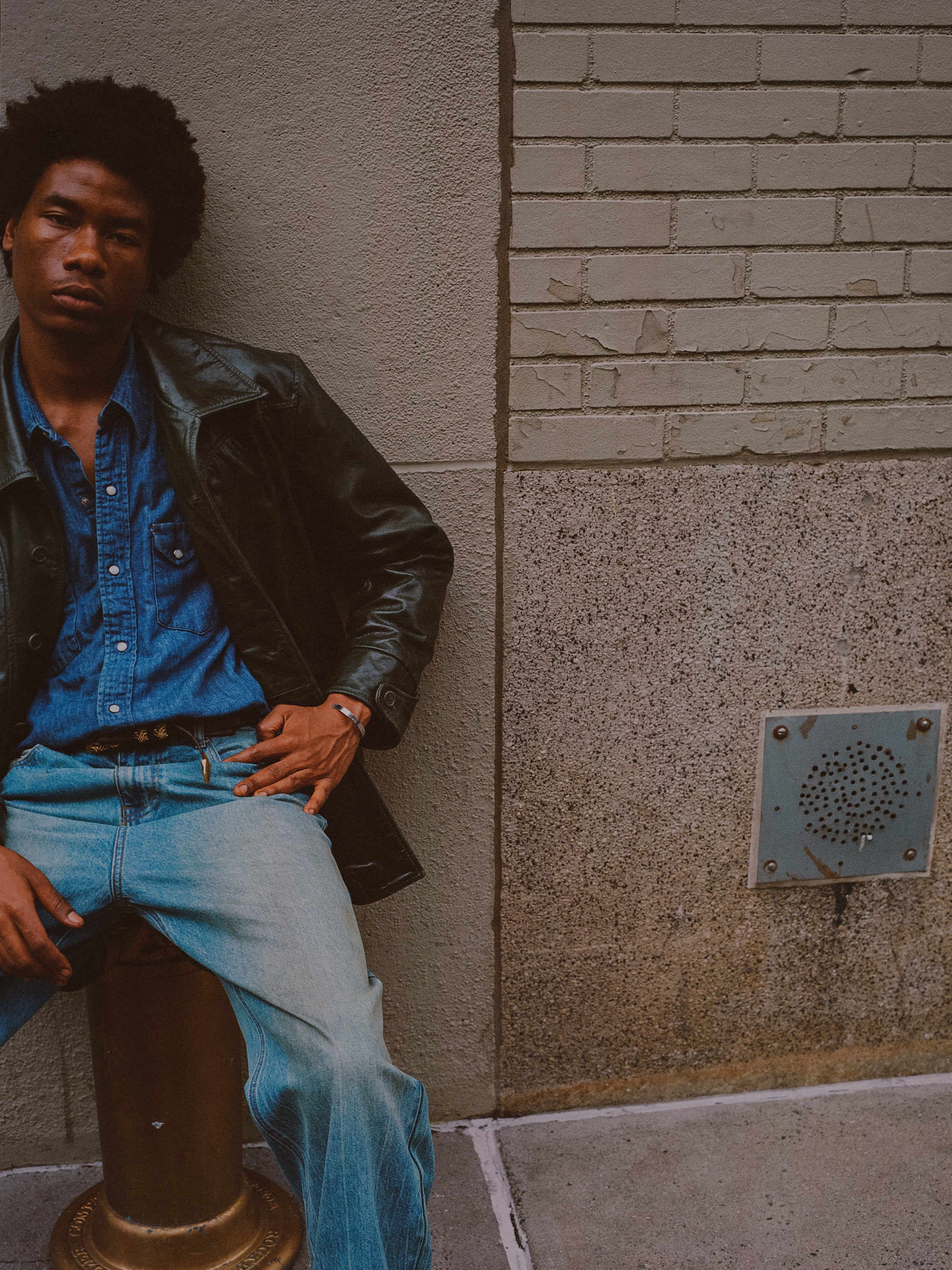 A man wearing an emerald leather jacket by FOUND and an emerald green denim outfit sits on a bollard against a concrete and brick wall.