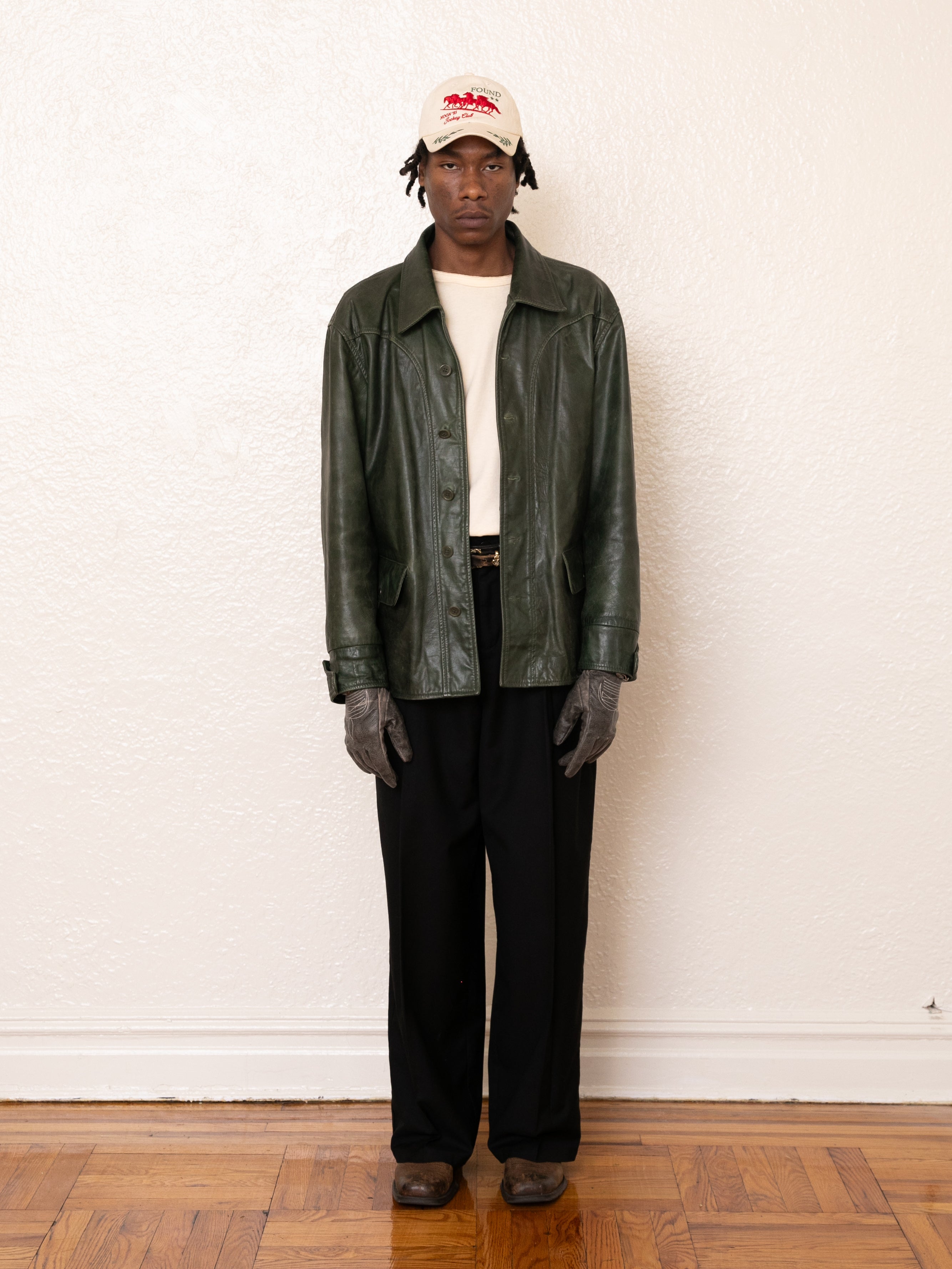 A person wearing an emerald leather jacket from FOUND, a white cap, gloves, a beige shirt, and black pants stands against a white wall on a wooden floor.