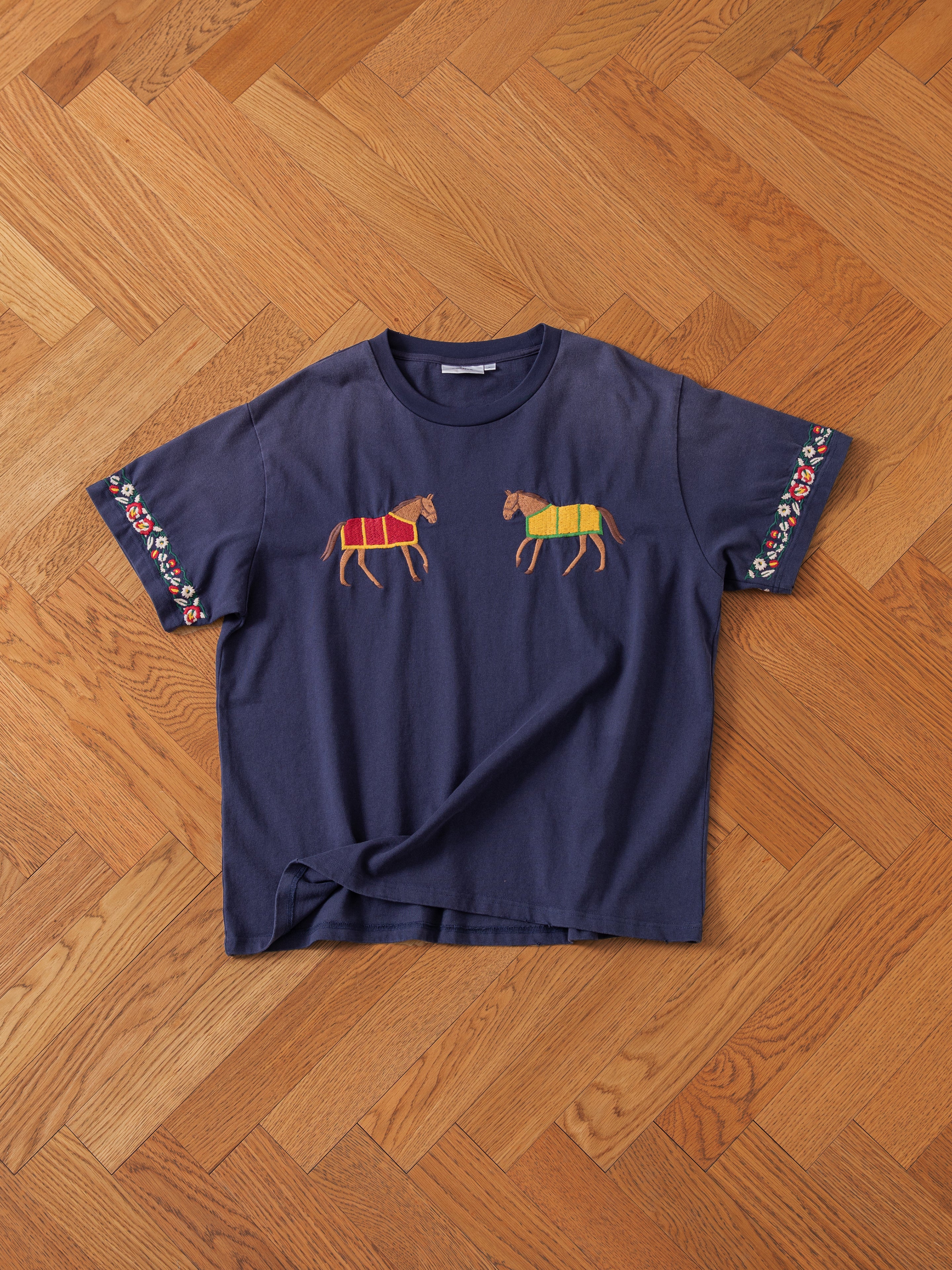 The Horse Equine Tee by FOUND, a navy blue t-shirt with embroidered horses in colorful blankets on the front and floral patterns along the sleeve hems, is laid flat on a wooden herringbone-pattern parquet floor.