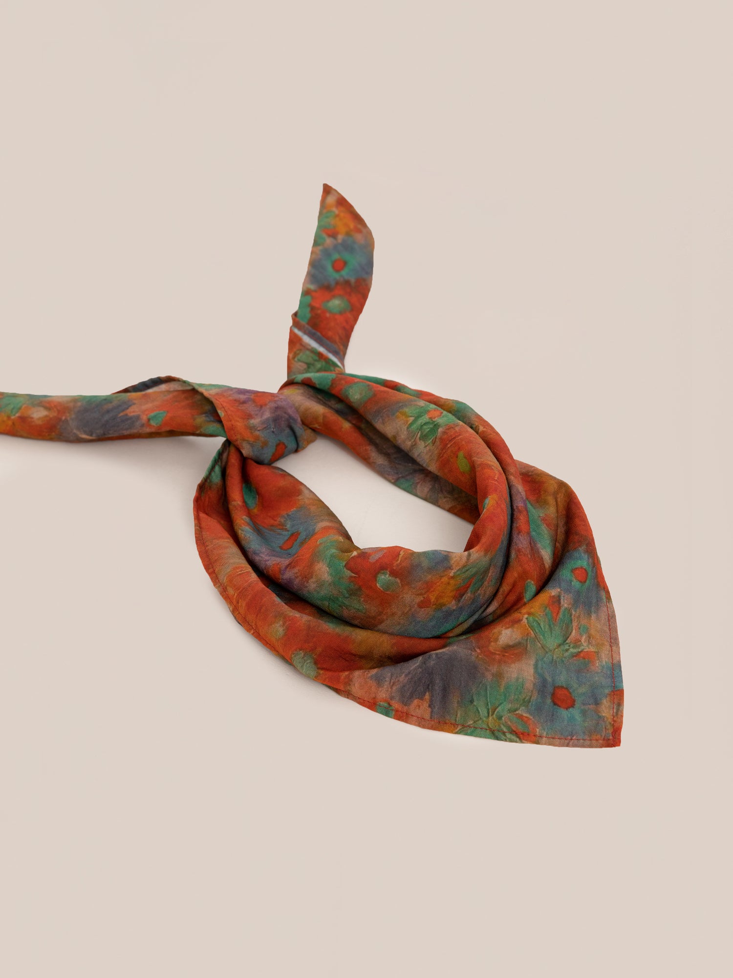 The Sepehr Bandana by FOUND, featuring a multicolored floral pattern with red, green, blue, and orange hues, is tied in a loose knot and laying against a beige background.