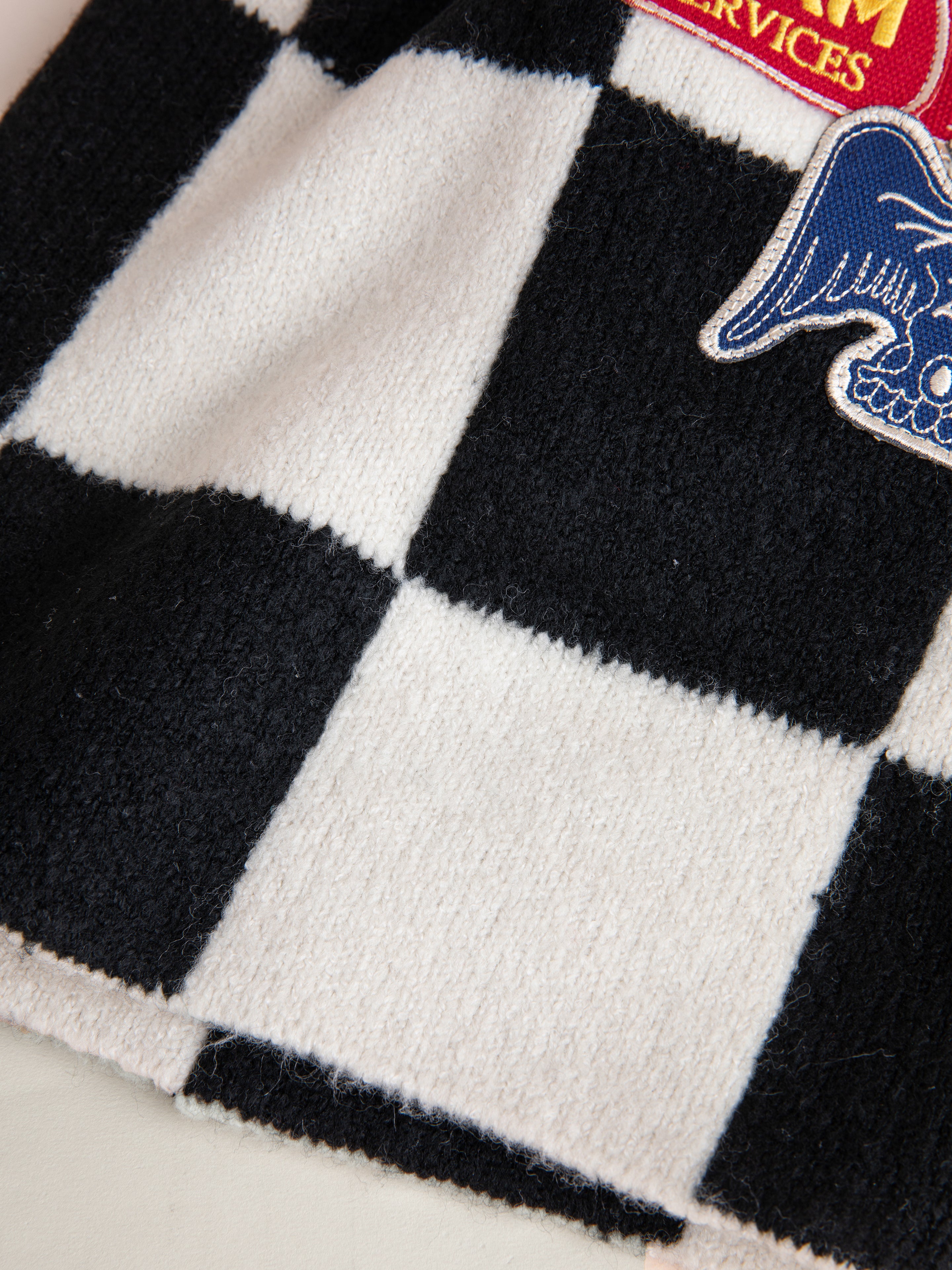 A close-up view of the fabric with a black and white checkered pattern, featuring embroidered patches — one in red and yellow, another with a blue and white winged skull design. The soft texture of the fabric is reminiscent of the FOUND Check Multi Patch Beanie.