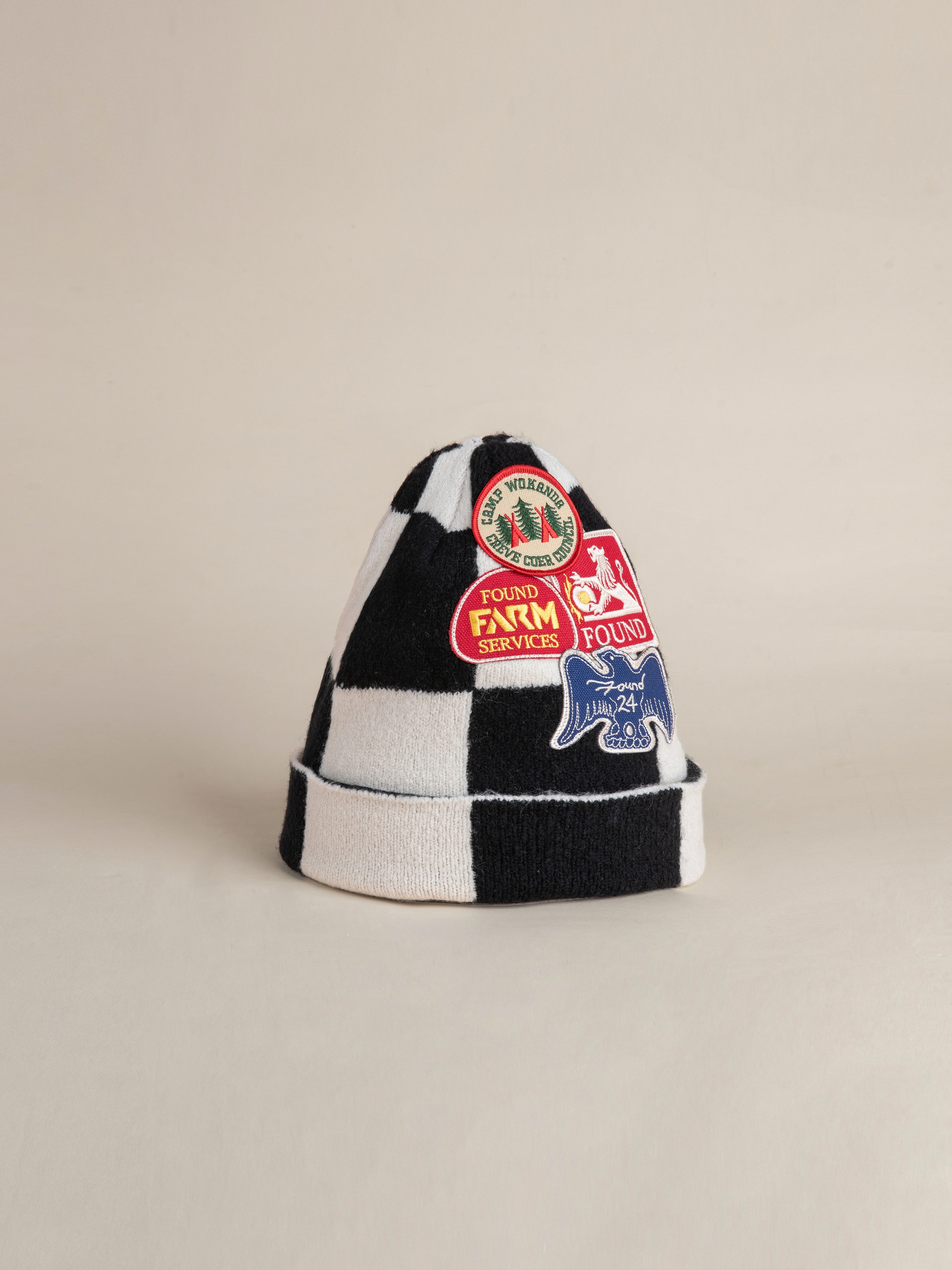 The Check Multi Patch Beanie by FOUND, featuring a black and white checkered knit design adorned with various colorful embroidered logos and emblems, is displayed against a plain background.