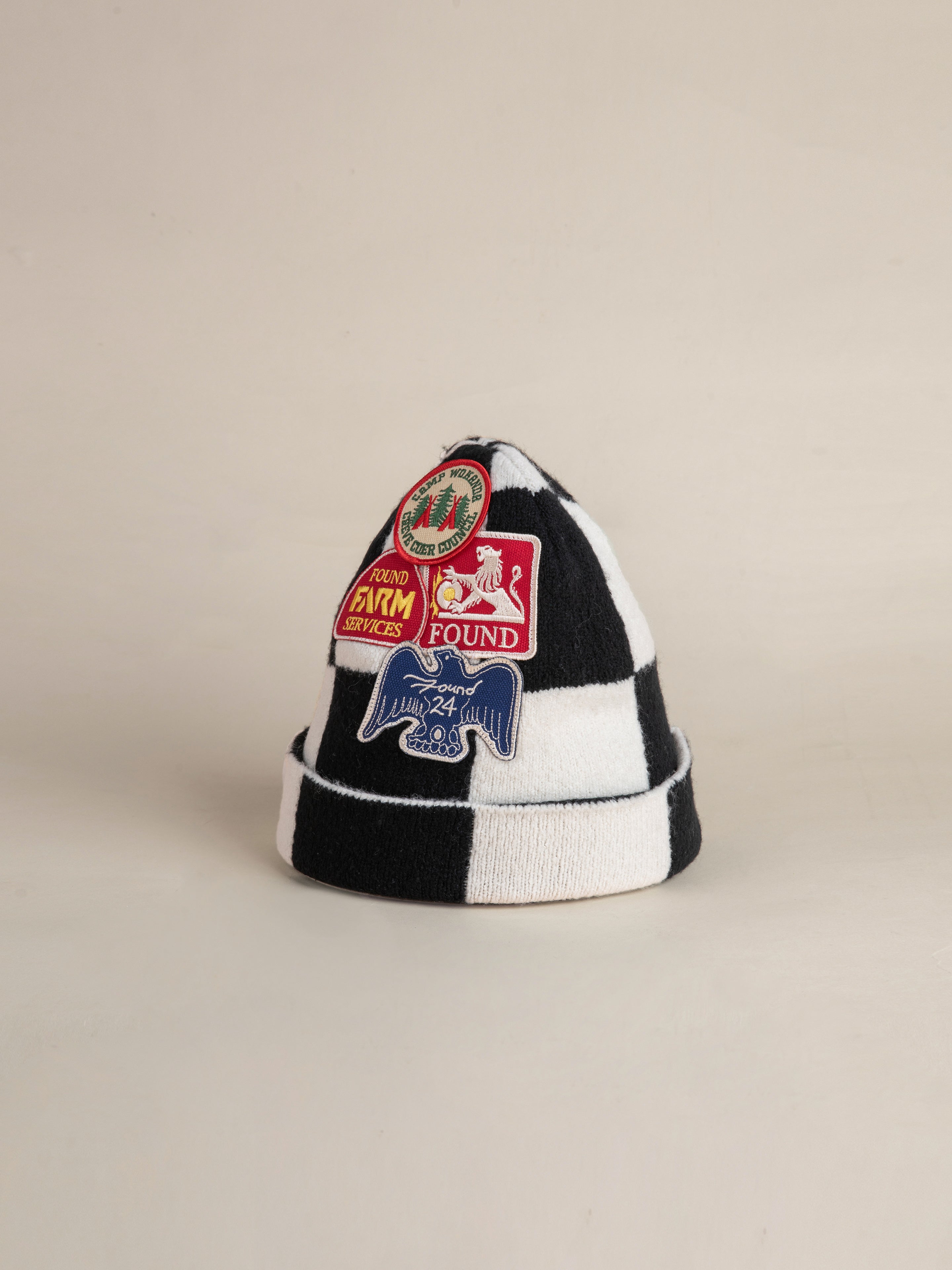 The FOUND Check Multi Patch Beanie boasts a striking black and white checkered pattern and is adorned with an array of colorful embroidered patches, featuring logos, text, and graphic designs.