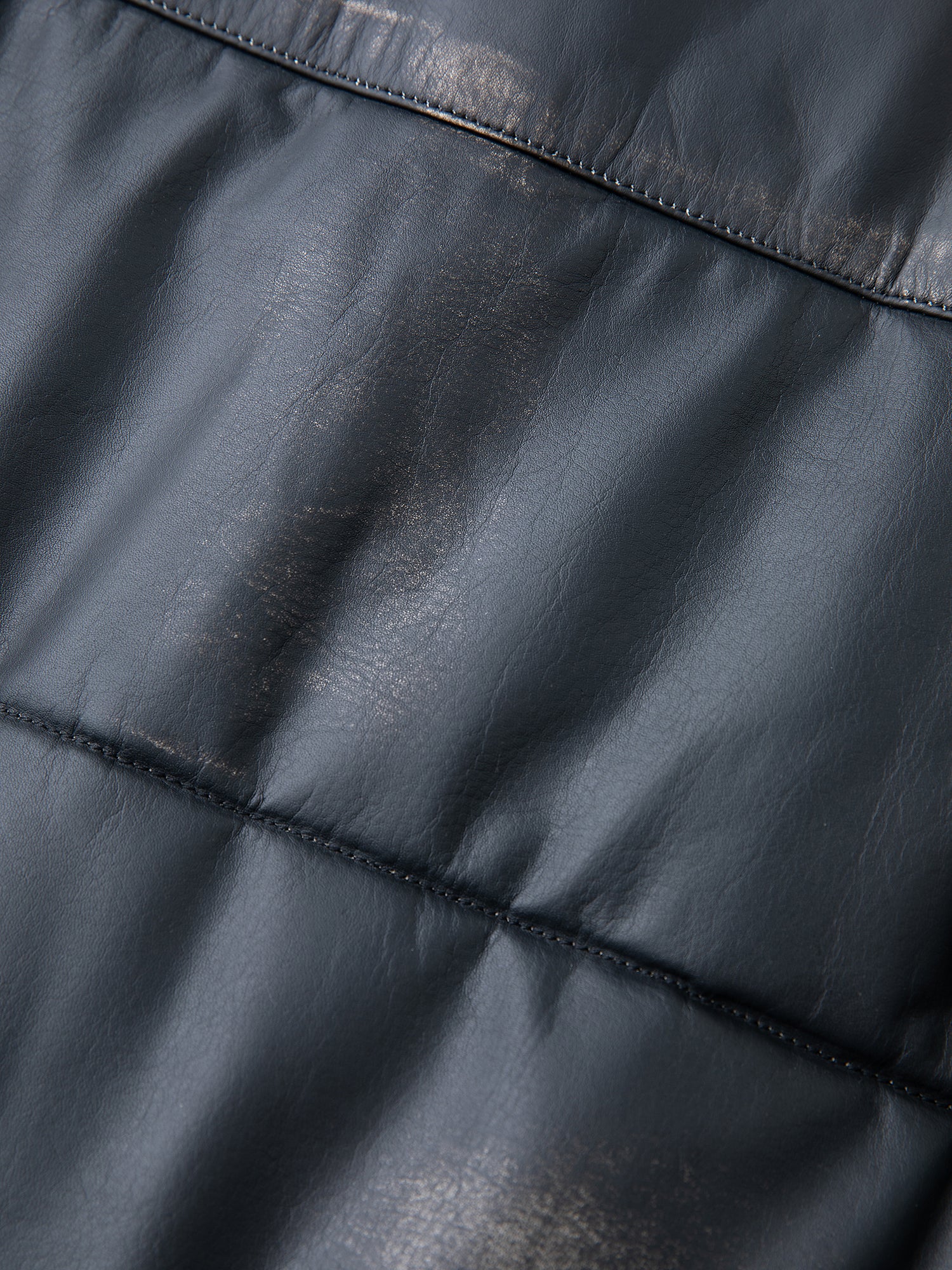 The close-up reveals the smooth, dark leather surface of our FOUND Leather Distressed Puffer Jacket, showcasing visible stitching, a slightly shiny finish, and natural creases and folds that highlight its genuine craftsmanship.