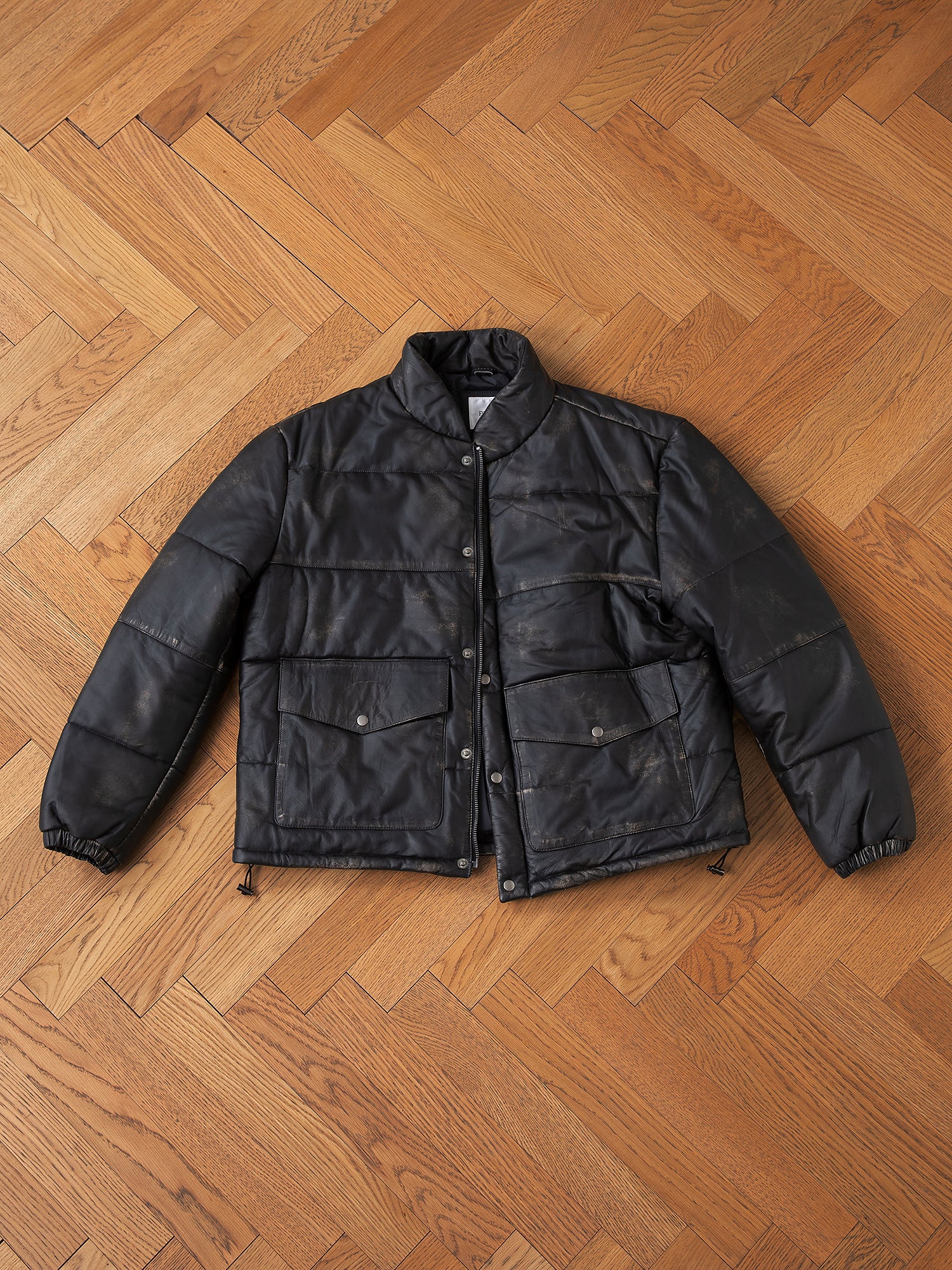 The Leather Distressed Puffer by FOUND is elegantly displayed on a herringbone-patterned wooden floor. This black padded jacket, made from ethically sourced materials, boasts a front button-up closure and large pockets, combining style with conscientious craftsmanship.