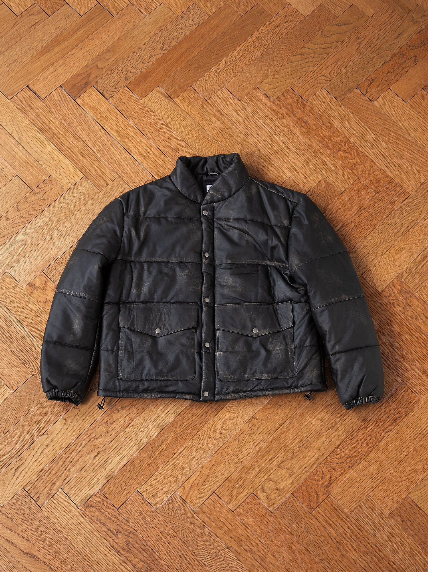 The FOUND Leather Distressed Puffer is crafted with ethically sourced materials and is displayed laid flat on a wooden herringbone-patterned floor. It boasts button and zipper closures, along with front pockets for added functionality.