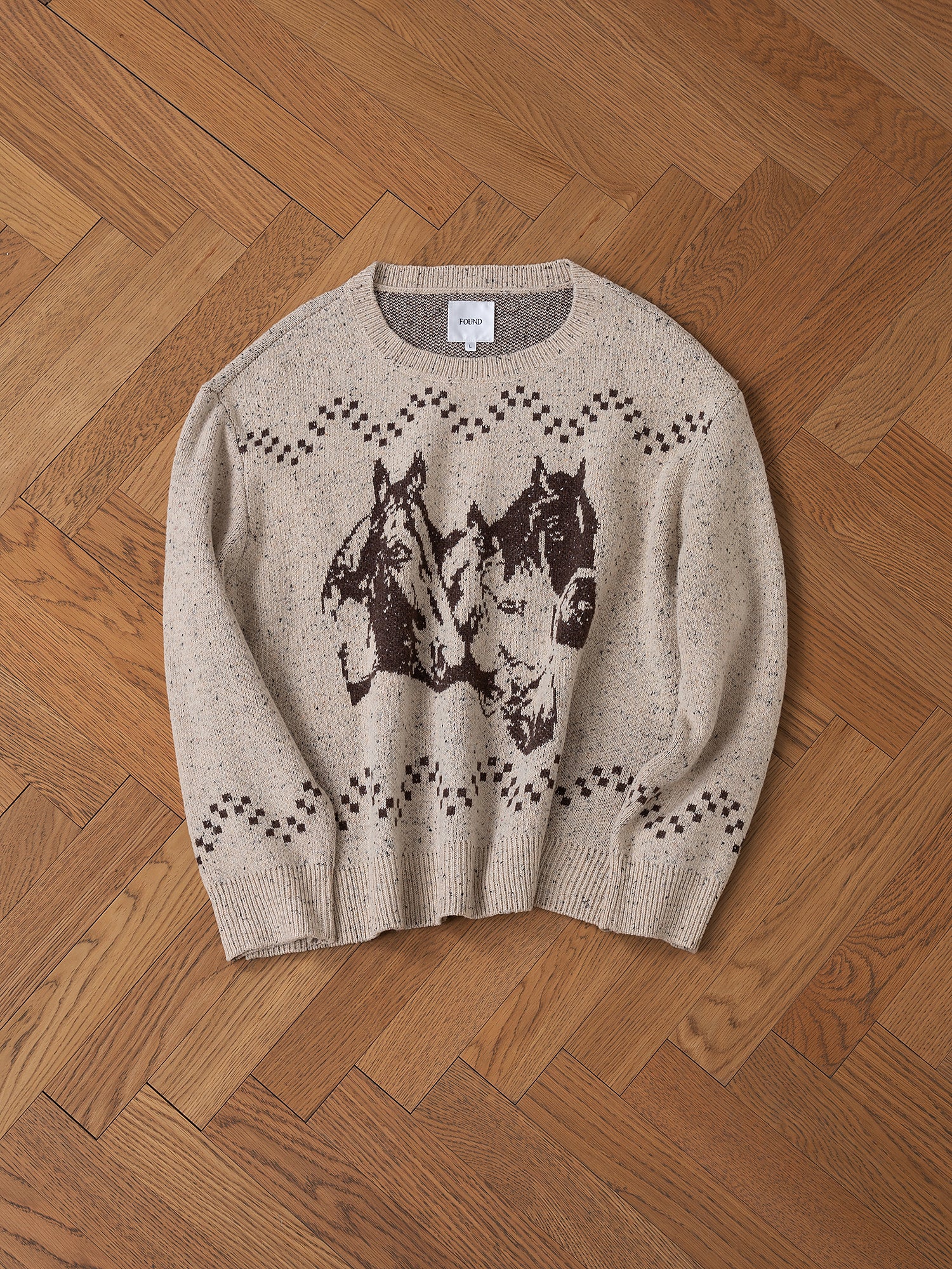 The Speckled Knit Horse Sweater from FOUND, made of a beige wool blend and featuring an oversized fit, showcases two brown jacquard horse faces and a checkered pattern, laid flat on a wooden herringbone floor.