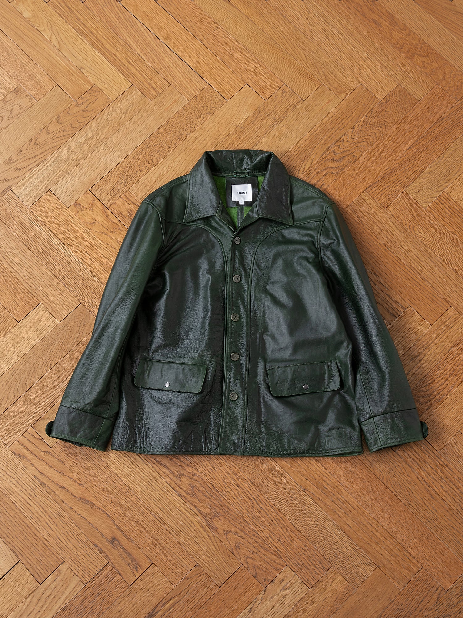 An Emerald Leather Jacket from FOUND, made with hand-distressed leather featuring buttons and two pockets, is laid flat on a herringbone parquet floor.