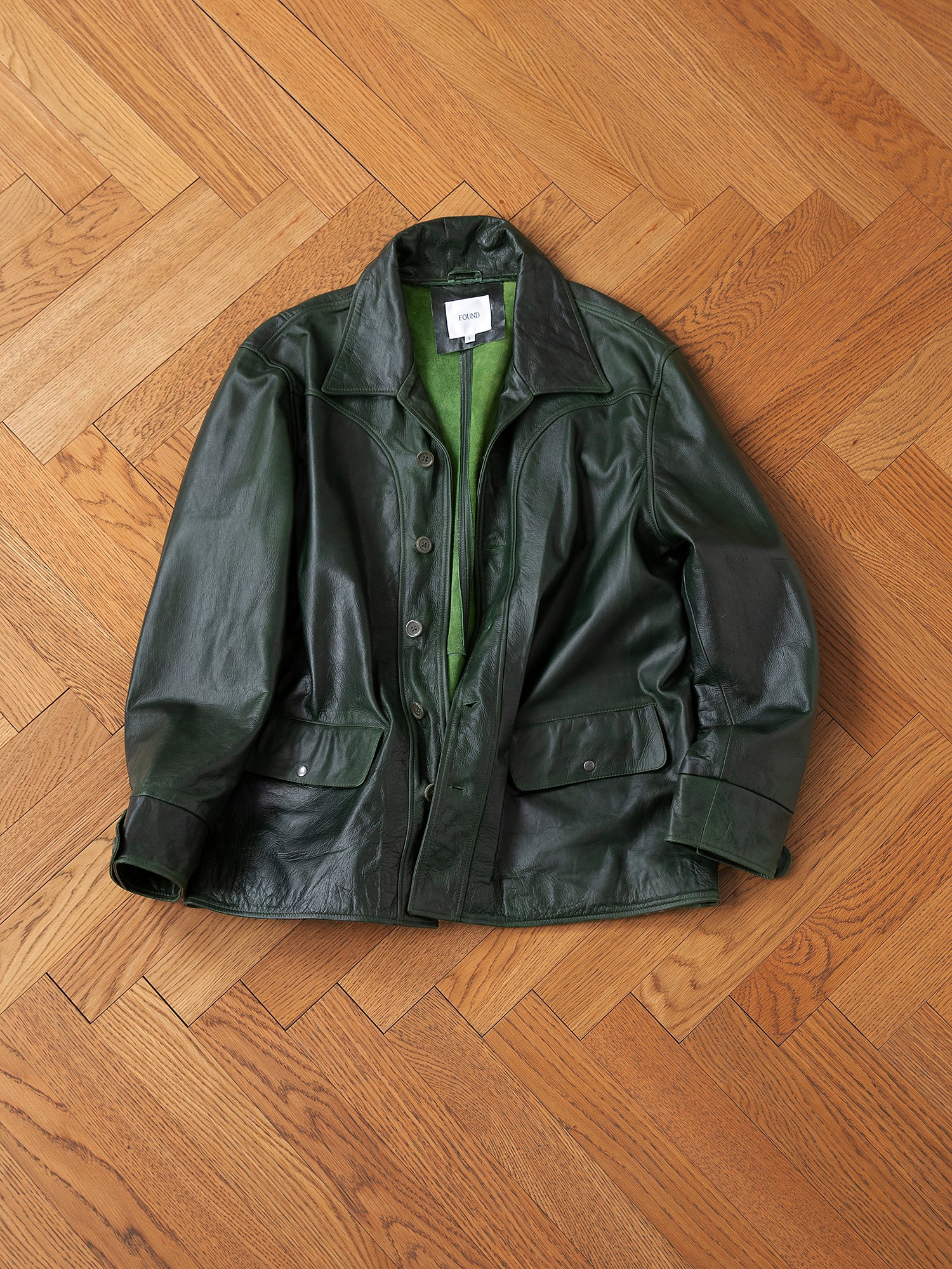 The Emerald Leather Jacket by FOUND, made of hand-distressed leather with a matching lining, elegantly rests flat on a wooden herringbone floor.