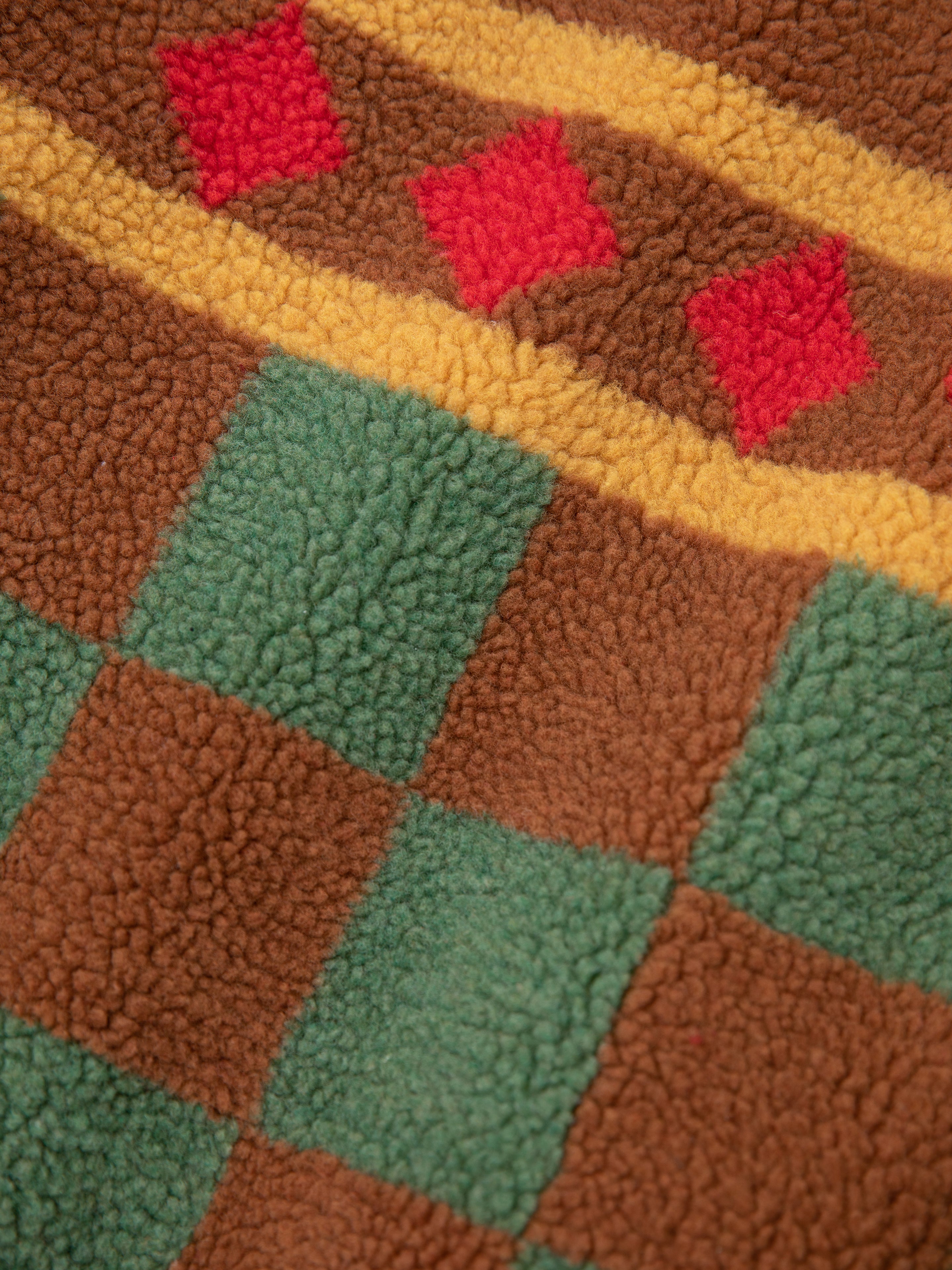 The FOUND Checkered Zip-Up Sherpa showcases a textured jacquard checkered design. It artfully combines green and brown squares with yellow line accents and red diamond shapes, offering an ideal blend of warmth and style.