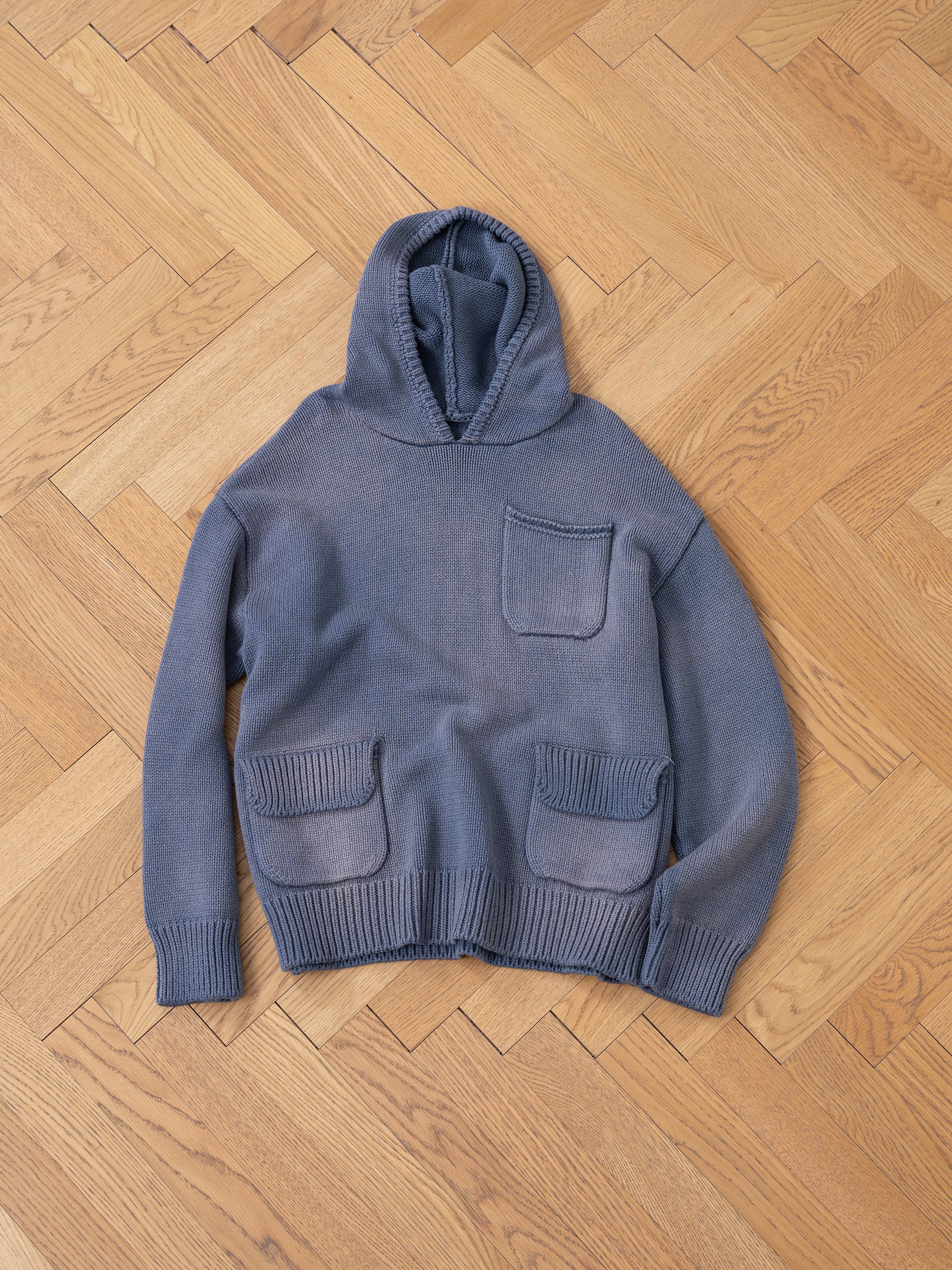 A Faded Knit Pocket Hoodie from FOUND, featuring an oversized cut in blue 100% cotton and three front pockets, is laid flat on a wooden parquet floor.