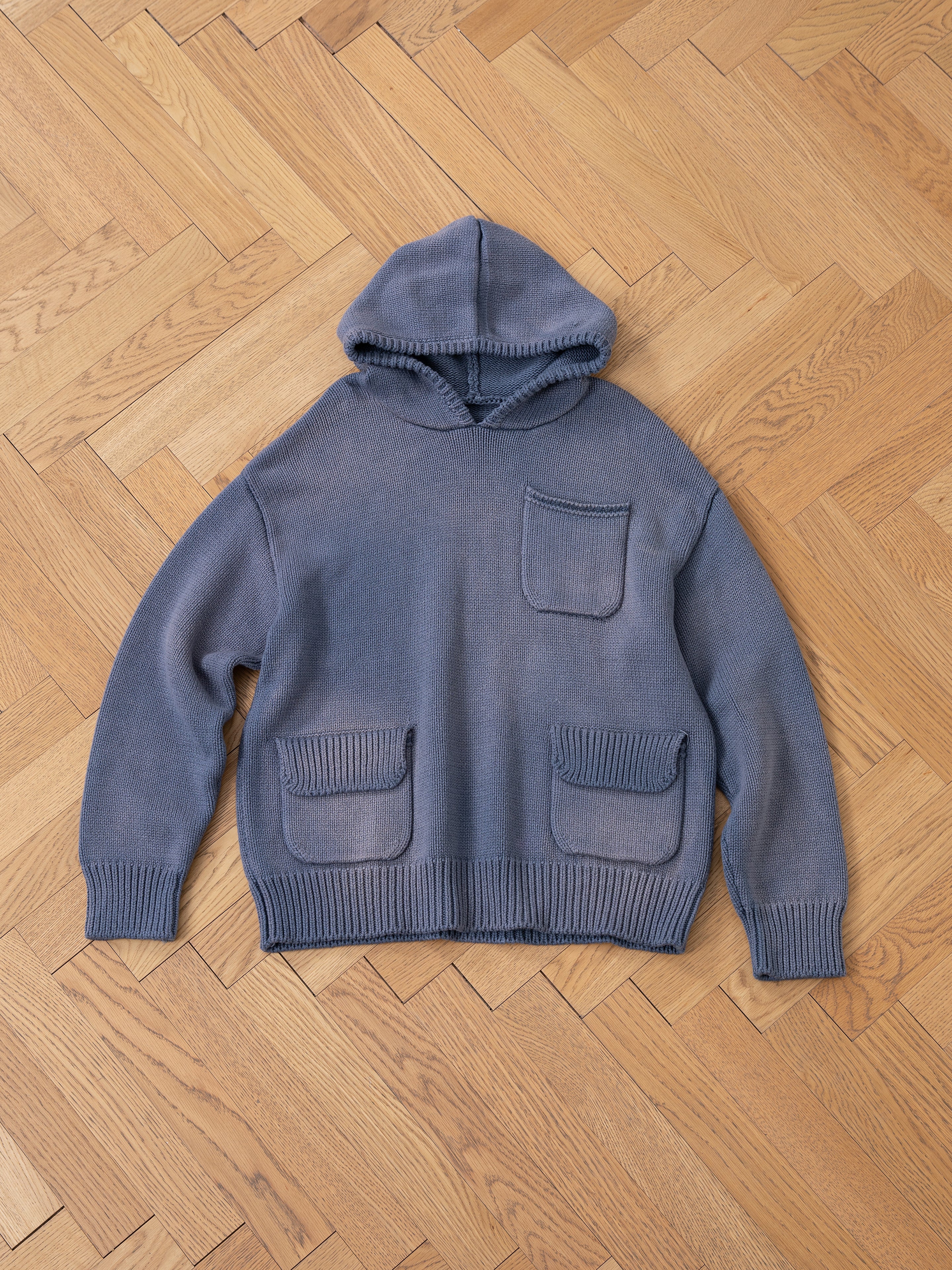 The Faded Knit Pocket Hoodie by FOUND, crafted from 100% cotton, features a blue oversized design with a front pocket and two spacious bottom pockets. Laid flat on a wooden parquet floor, it exudes cozy sophistication.