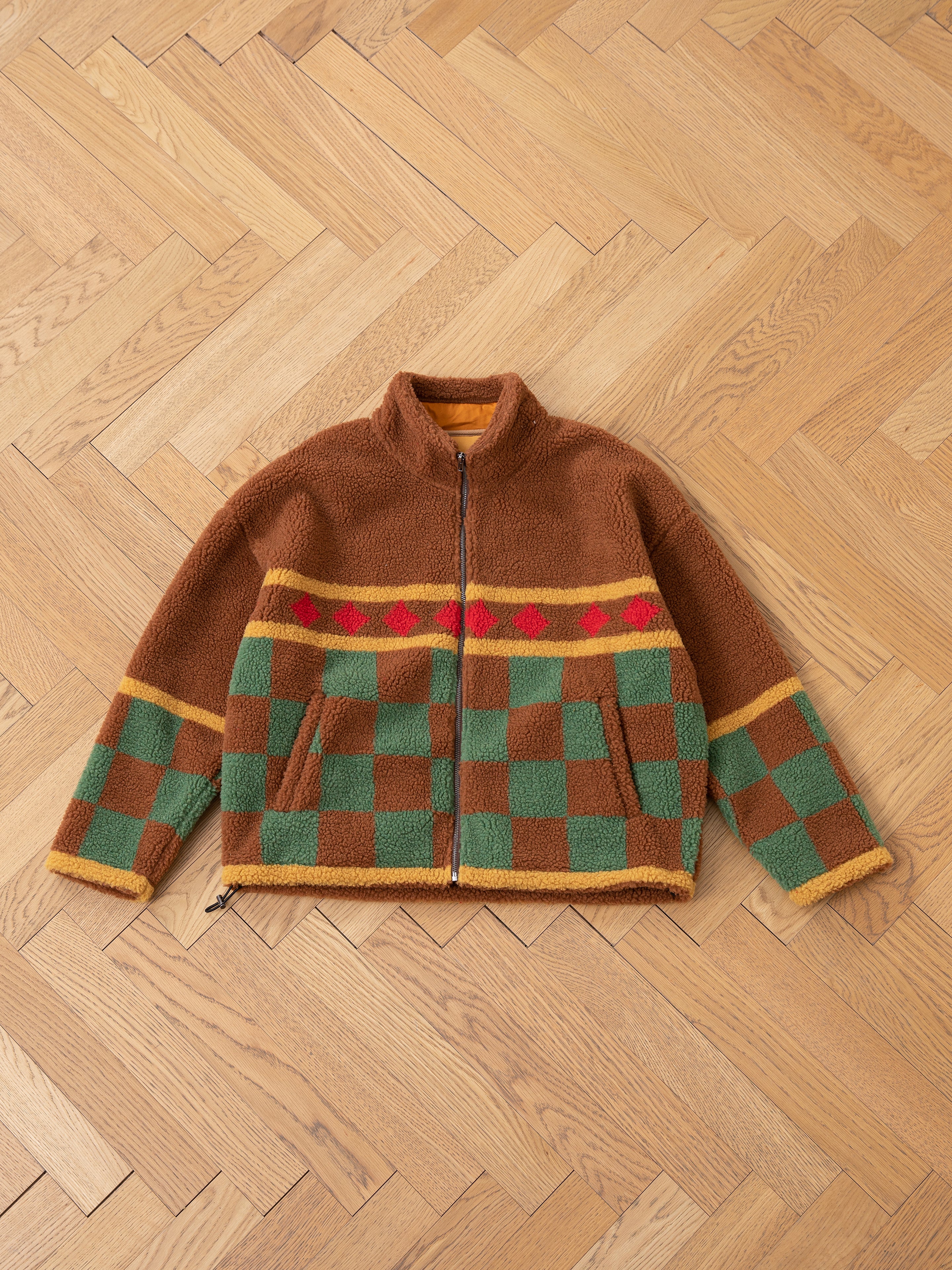 Checkered Zip-Up Sherpa by FOUND, featuring a brown color with red, green, and yellow jacquard checkered design, displayed flat on a wooden herringbone floor.