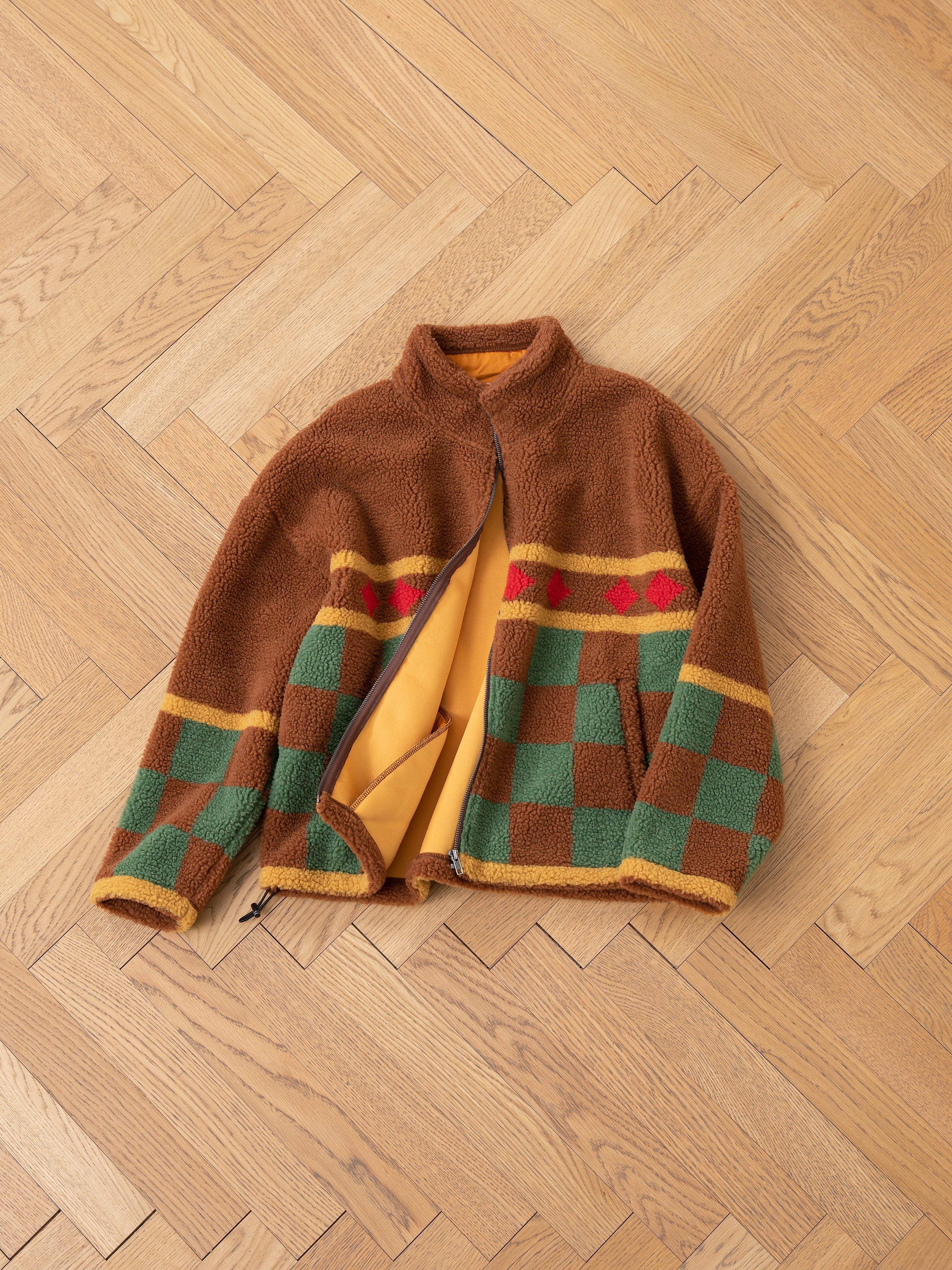 The Checkered Zip-Up Sherpa by FOUND is an oversized fleece jacket with a brown base, showcasing a jacquard checkered design in green and yellow with red accents. It is displayed open on a wooden herringbone floor.