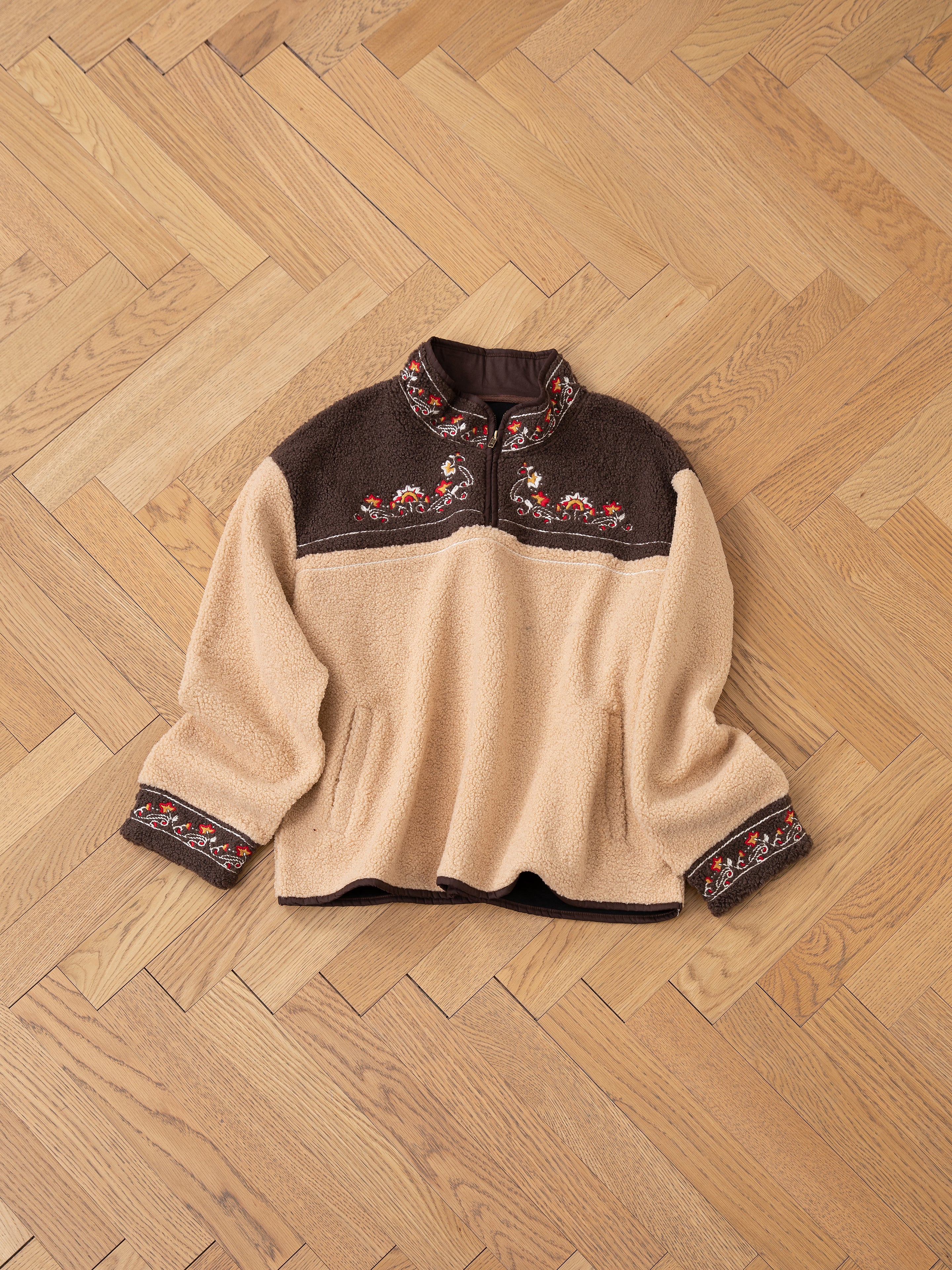 A Pahari Floral Embroidered Sherpa Pullover by FOUND, in beige and brown, showcases exquisite Pahari embroidery on the collar and cuffs, elegantly displayed on a light wood herringbone floor.