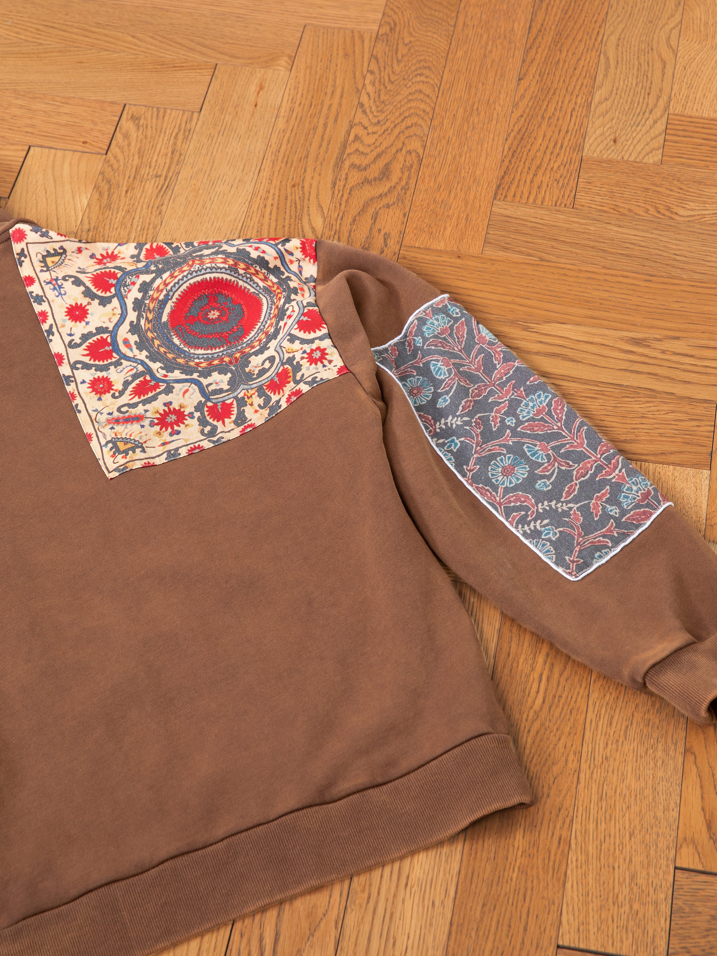 The Tapestry Patchwork Zip Up Hoodie by FOUND, designed in an oversized fit and featuring colorful floral patches on the shoulder and sleeve reminiscent of South Asian tapestry, is beautifully laid on a wooden floor.