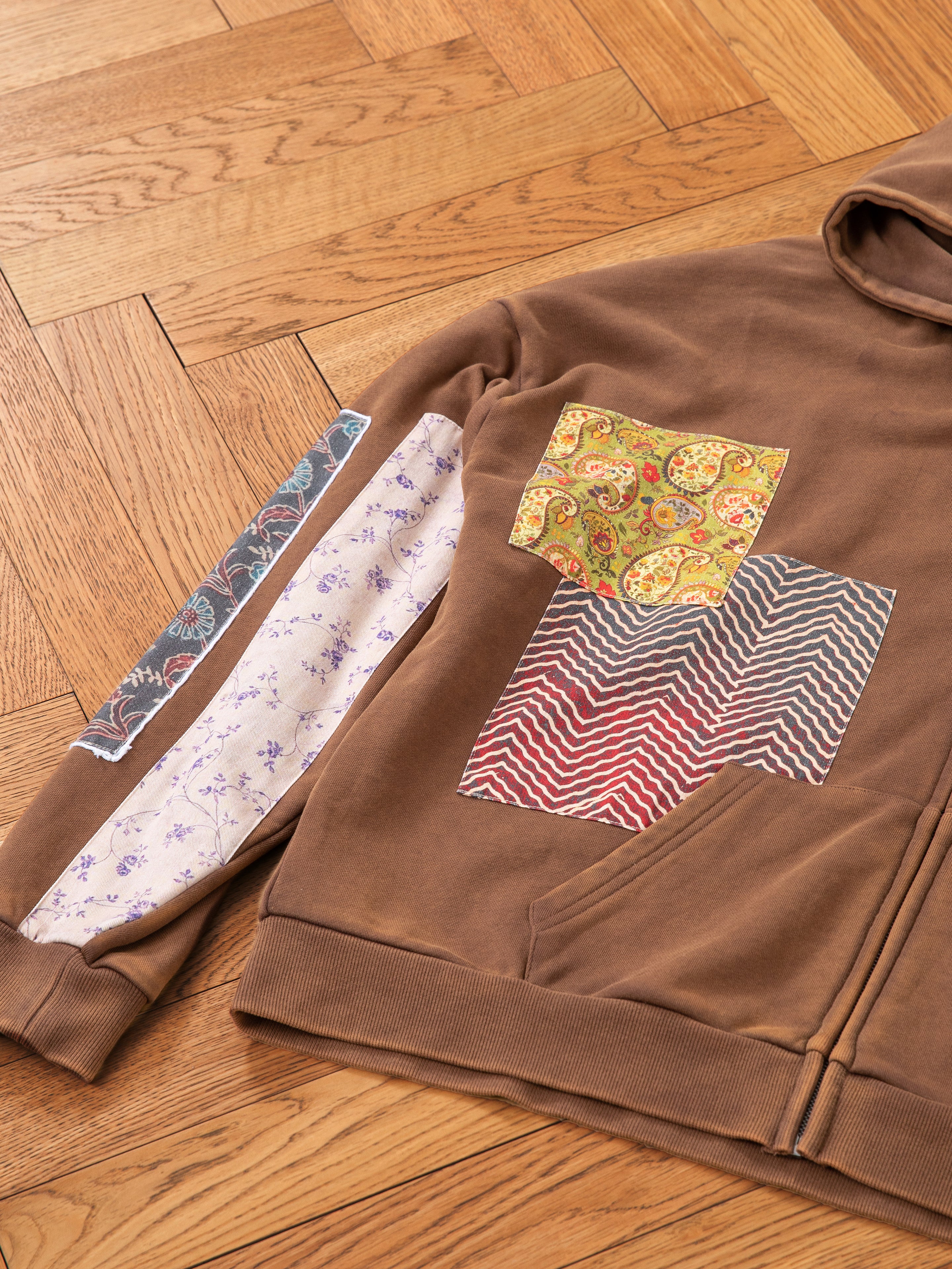 The Tapestry Patchwork Zip Up Hoodie by FOUND showcases colorful designs on the sleeves and front, featuring paisley, floral, and wavy patterns reminiscent of South Asian tapestry. This oversized fit piece is beautifully displayed on a wooden floor.