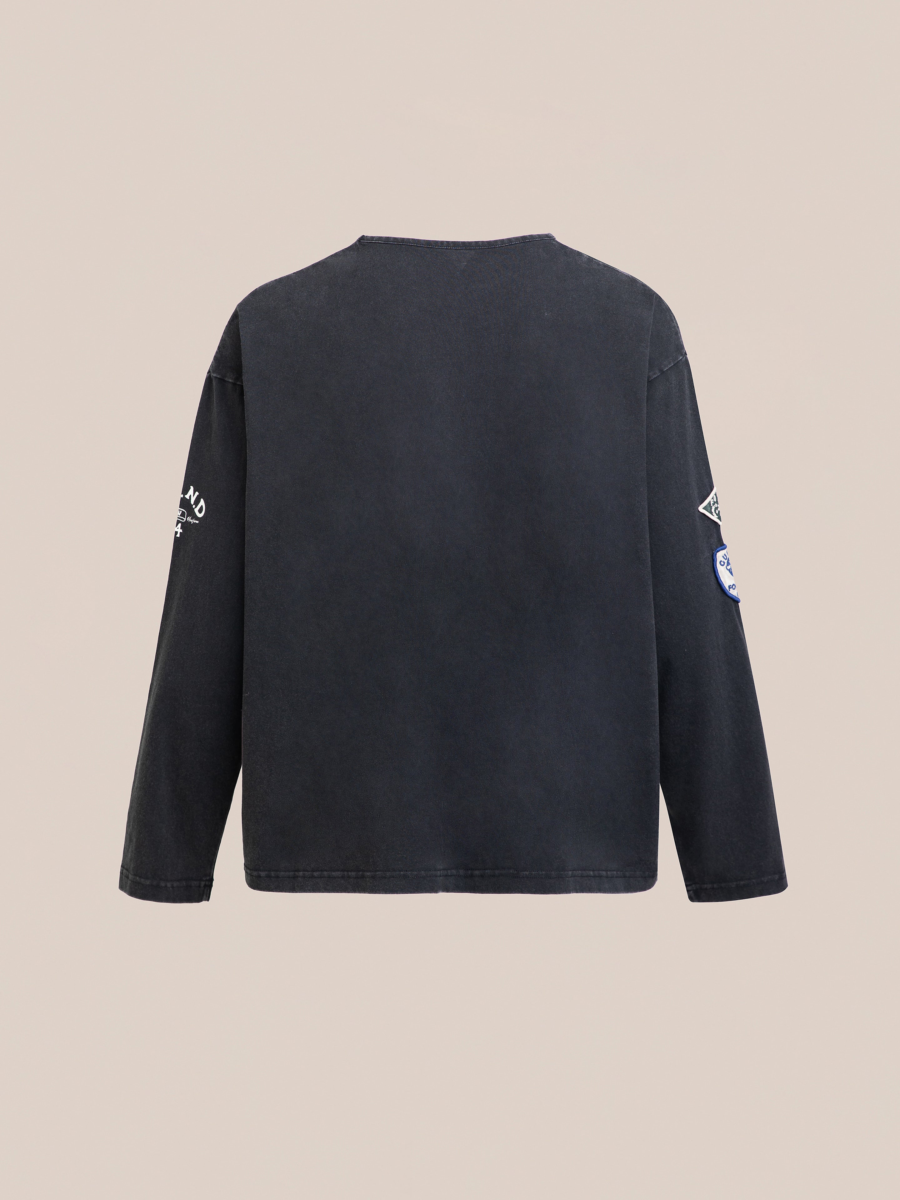 Back view of the Studded LS Shirt by FOUND in faded black with graphic patches on the sleeves, displayed against a plain beige background.