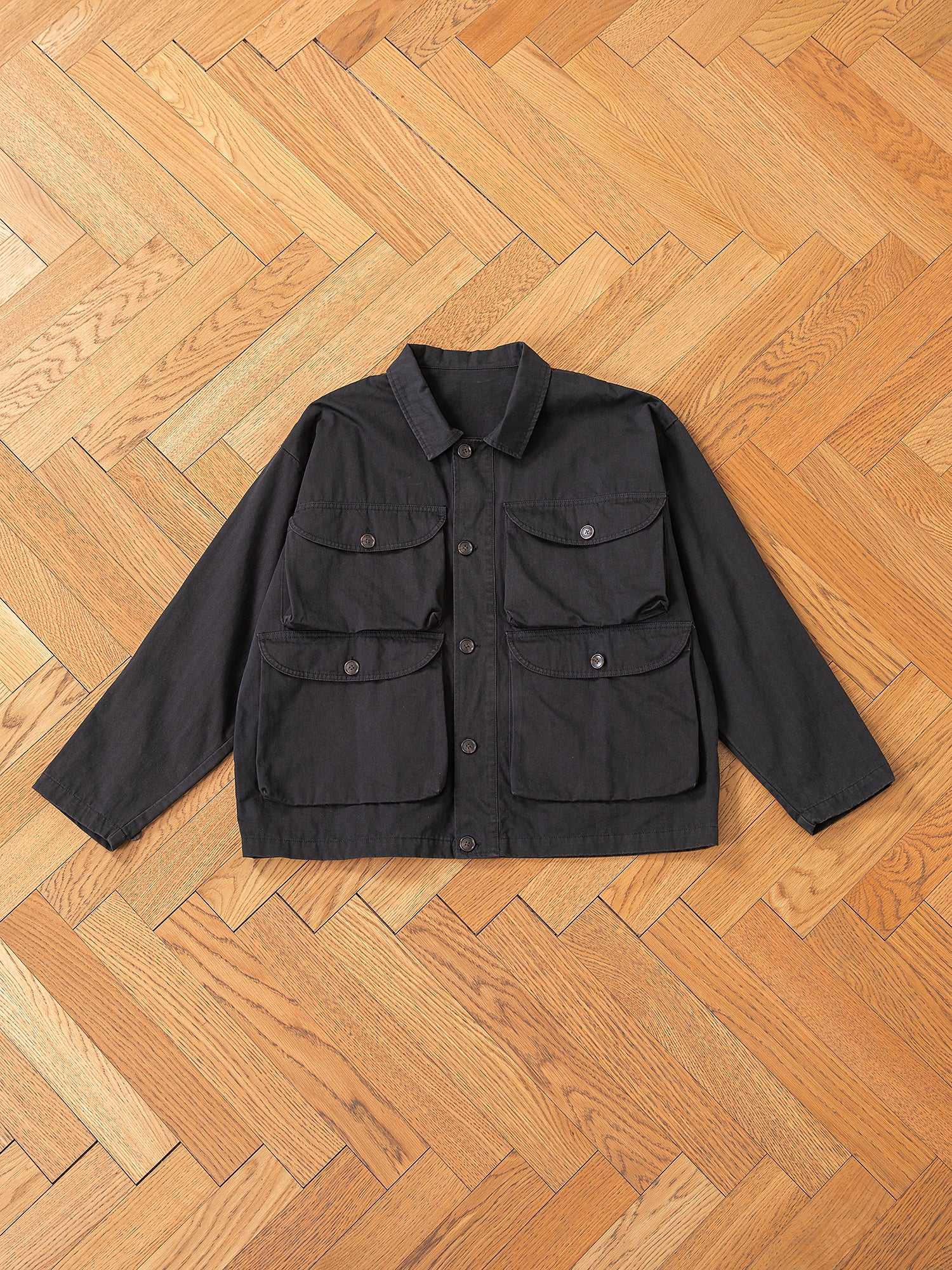 A FOUND Utility Herringbone Cargo Jacket in classic black, featuring a vintage design with four pockets, is laid flat on a wooden herringbone floor.