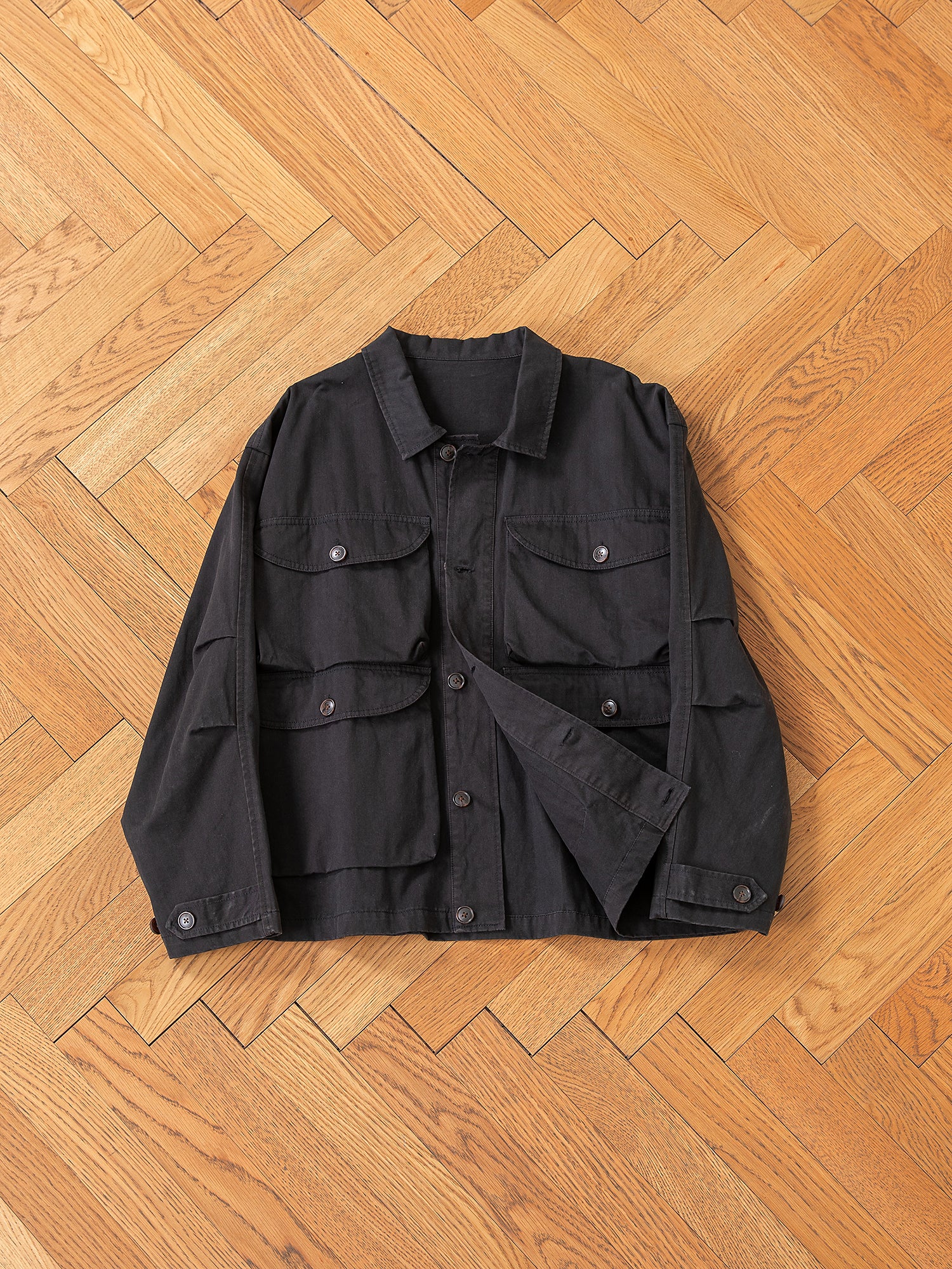 The FOUND Utility Herringbone Cargo Jacket offers a vintage appeal with its button closure and four front pockets, elegantly showcased on a wooden herringbone floor.