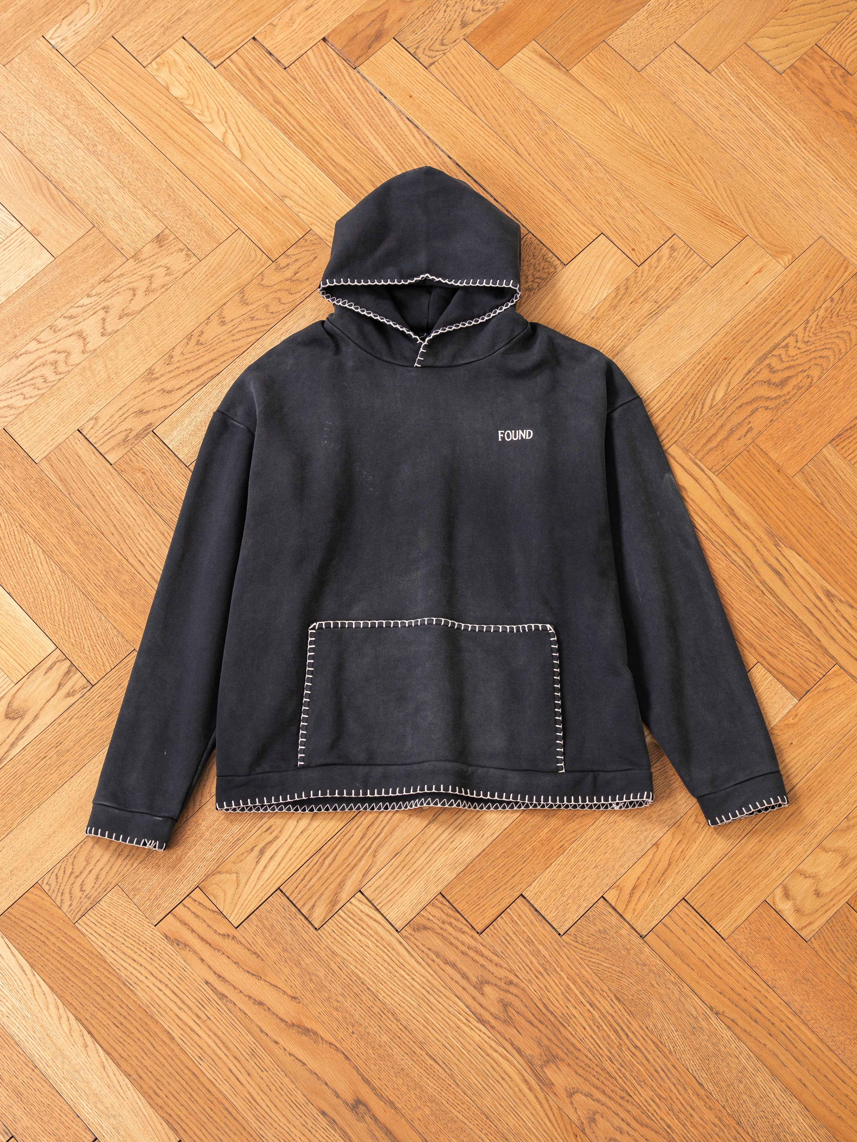 A FOUND hoodie, officially named the Stitched Border Distressed Logo Hoodie, made from French Terry Cotton in Vintage Black, is elegantly designed with a stitched border and features the brand's logo on the front. This stylish piece is showcased on a wooden herringbone floor.