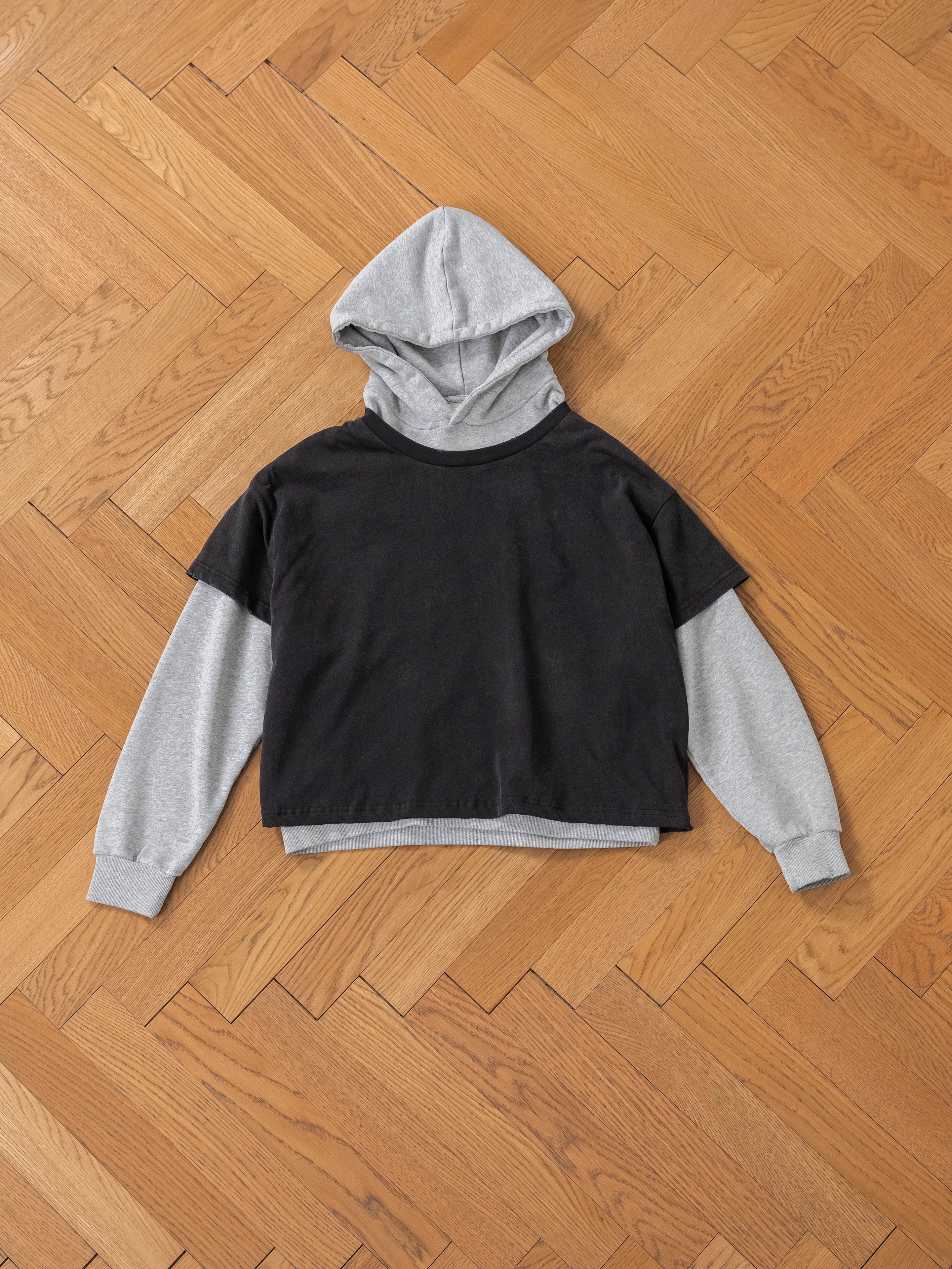 A stylish gray Double Layer Hoodie by Found, made from French terry cotton and featuring a black short-sleeved T-shirt overlay, is elegantly placed on a wooden herringbone-pattern floor.