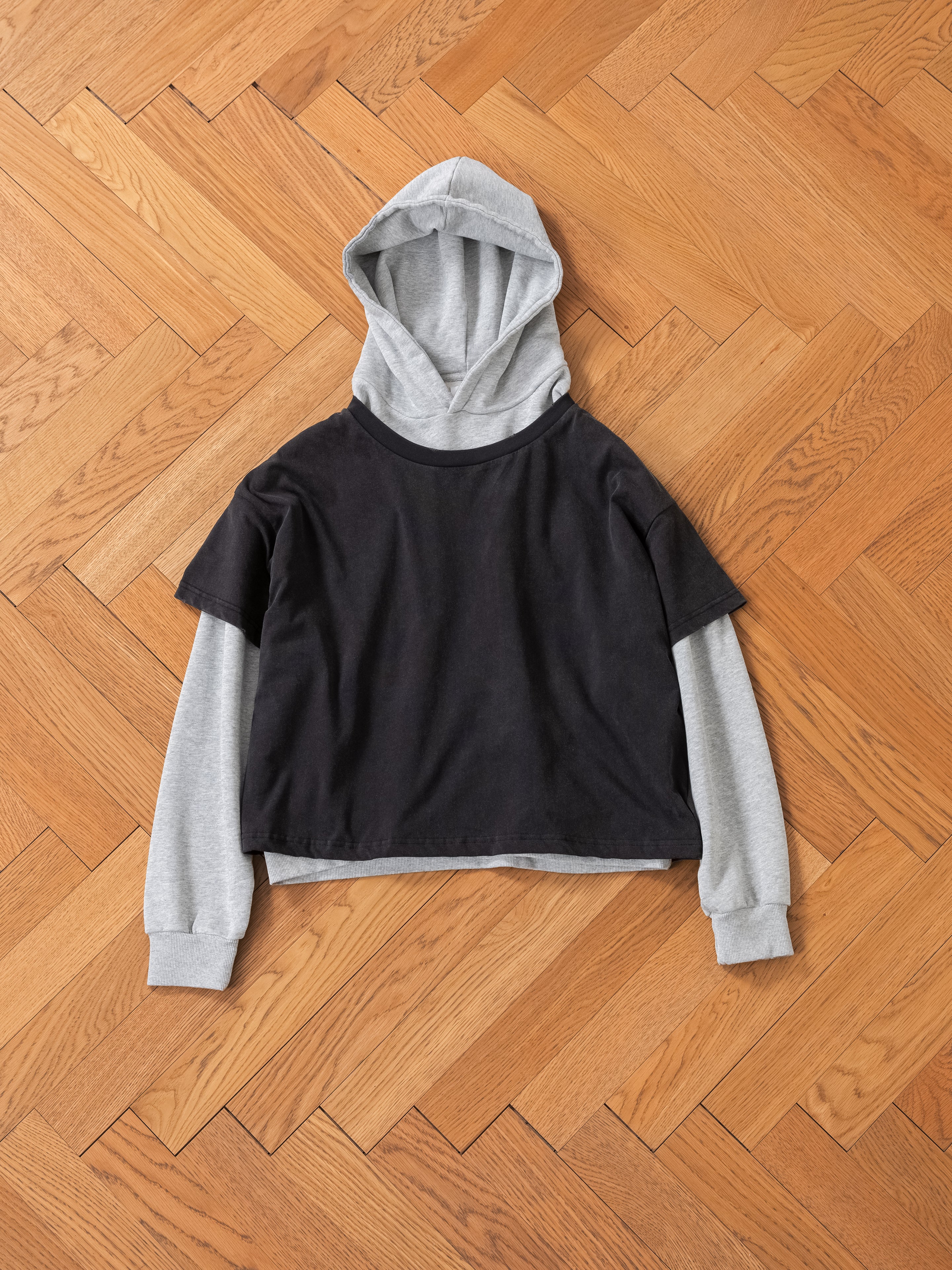 The Found Double Layer Hoodie, black with gray long sleeves and hood, crafted from French terry cotton, laid flat on a herringbone hardwood floor, captures a vintage look.