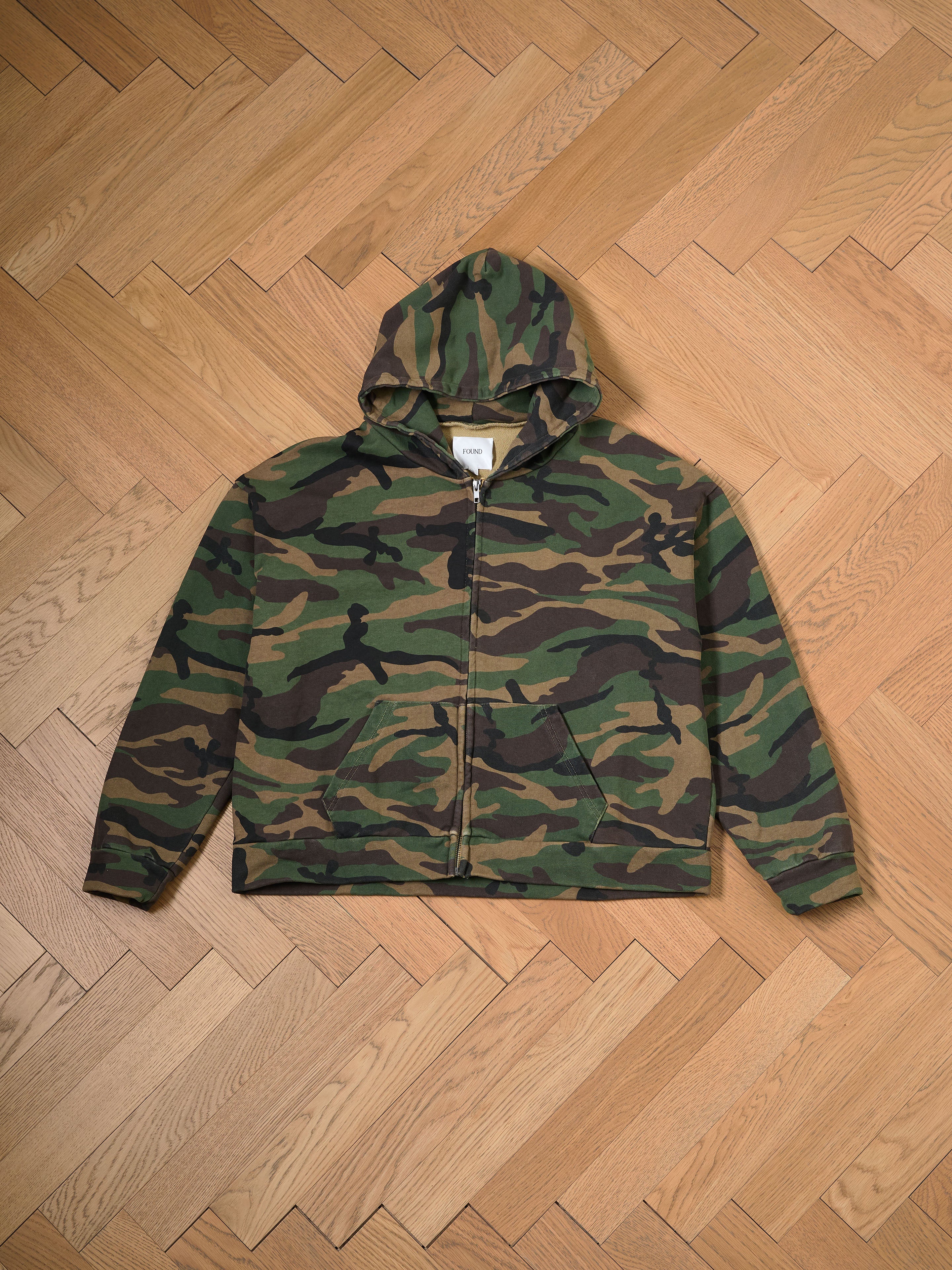 FOUND's Washed Camouflage Zip Hoodie, made of soft French terry cotton, showcases a stylish camouflage print and a practical front zipper, beautifully presented on a wooden herringbone floor.